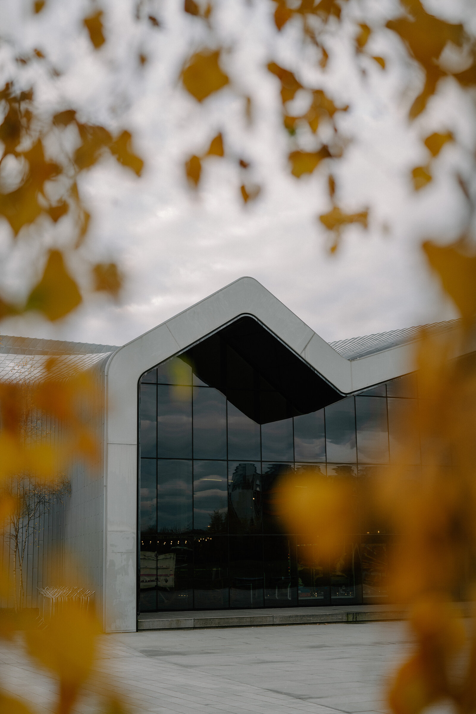 A close-up view of a zigzag-roofed museum building's white ribbed panels and dark glass frames a cloudy sky, autumn leaves blurred in the foreground.