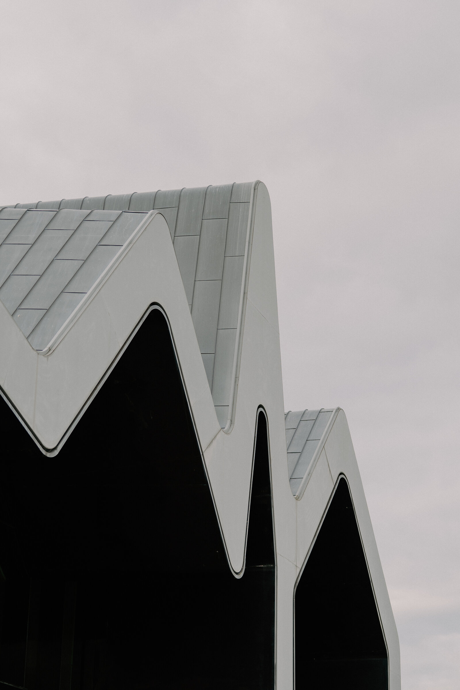 The zigzag rooftops of a modern museum building rise against an overcast sky, dark glass panels filling the triangular gables.