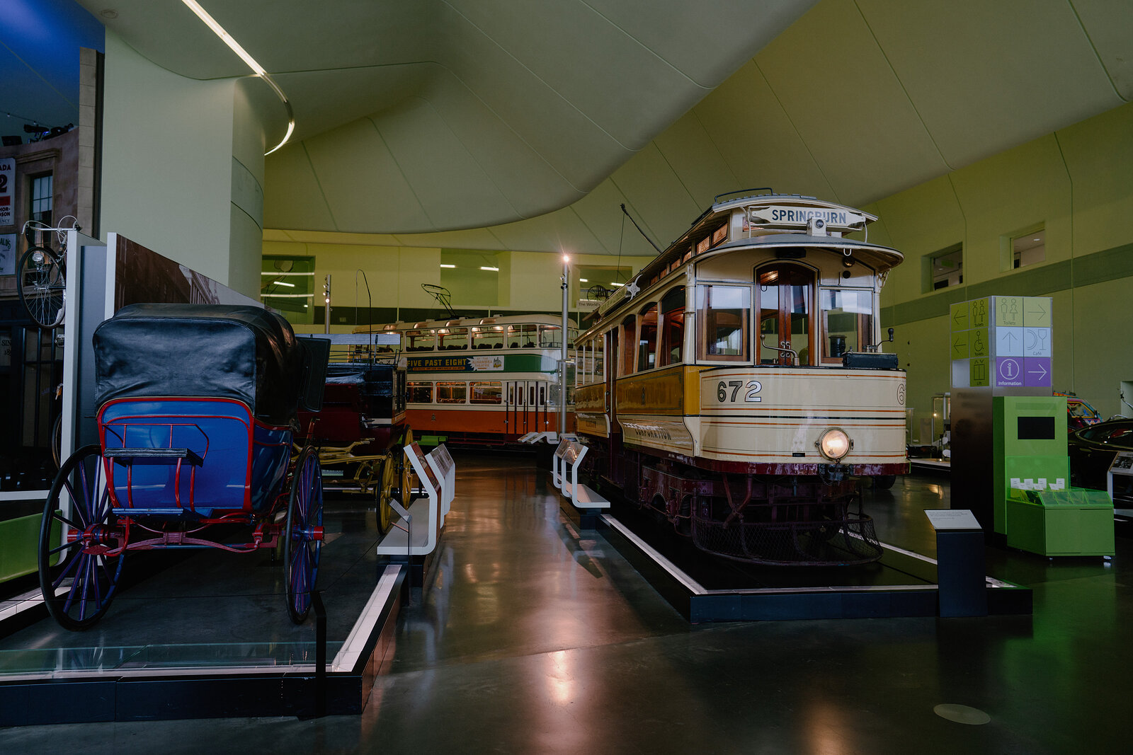 A museum gallery displays vintage horse-drawn carriages and tramcars, one numbered '672', beneath a curved ceiling.