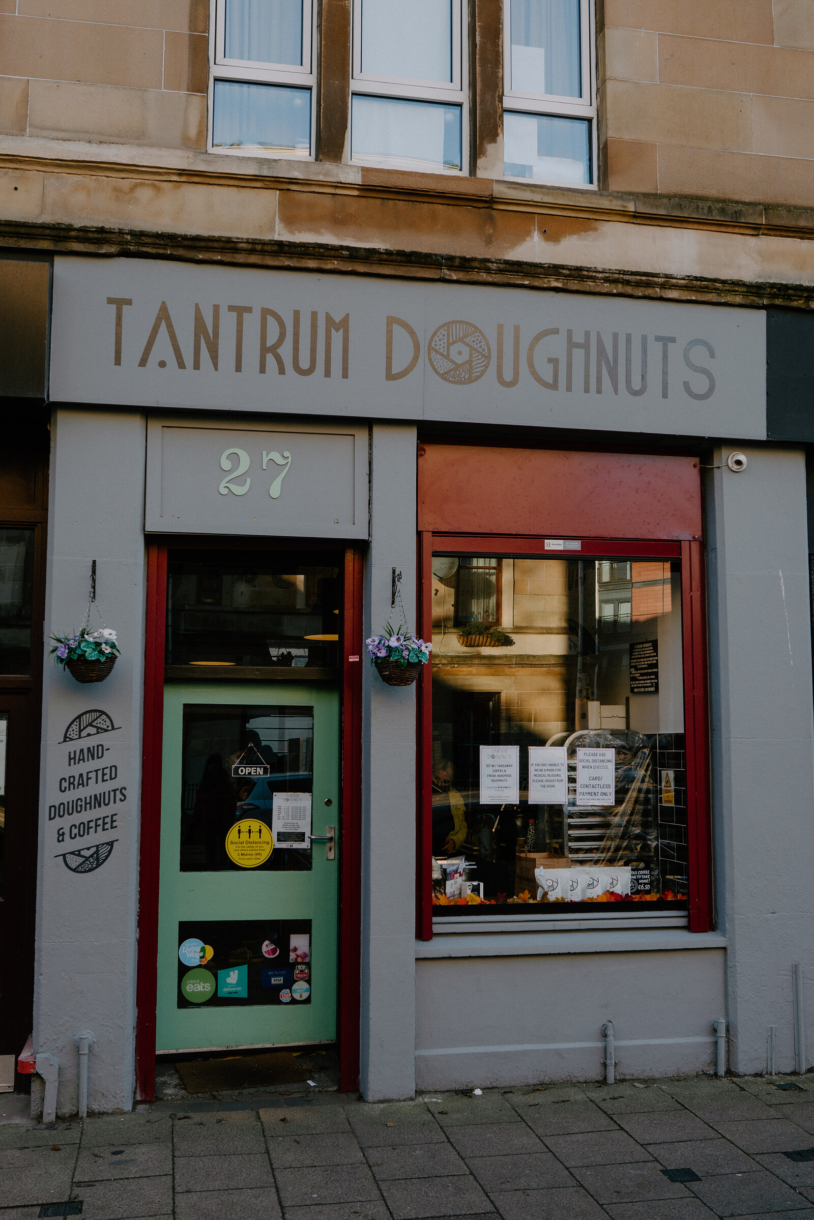 A grey shopfront carries a sign reading 'Tantrum Doughnuts' above a green door and red-framed window, hanging plant baskets on either side.