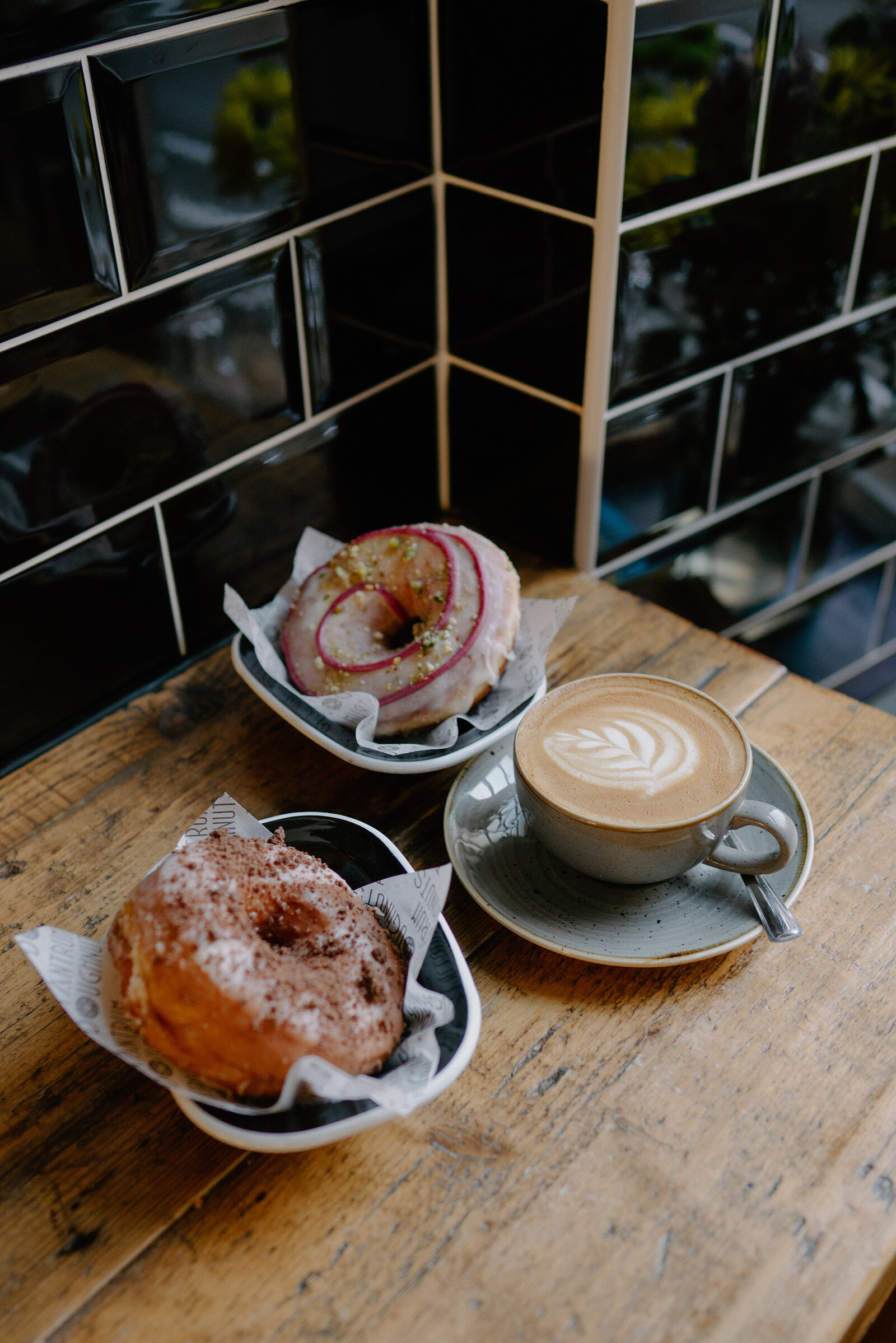 Two glazed doughnuts and a latte with leaf-pattern foam sit on small plates on a wooden table beside black-tiled walls.