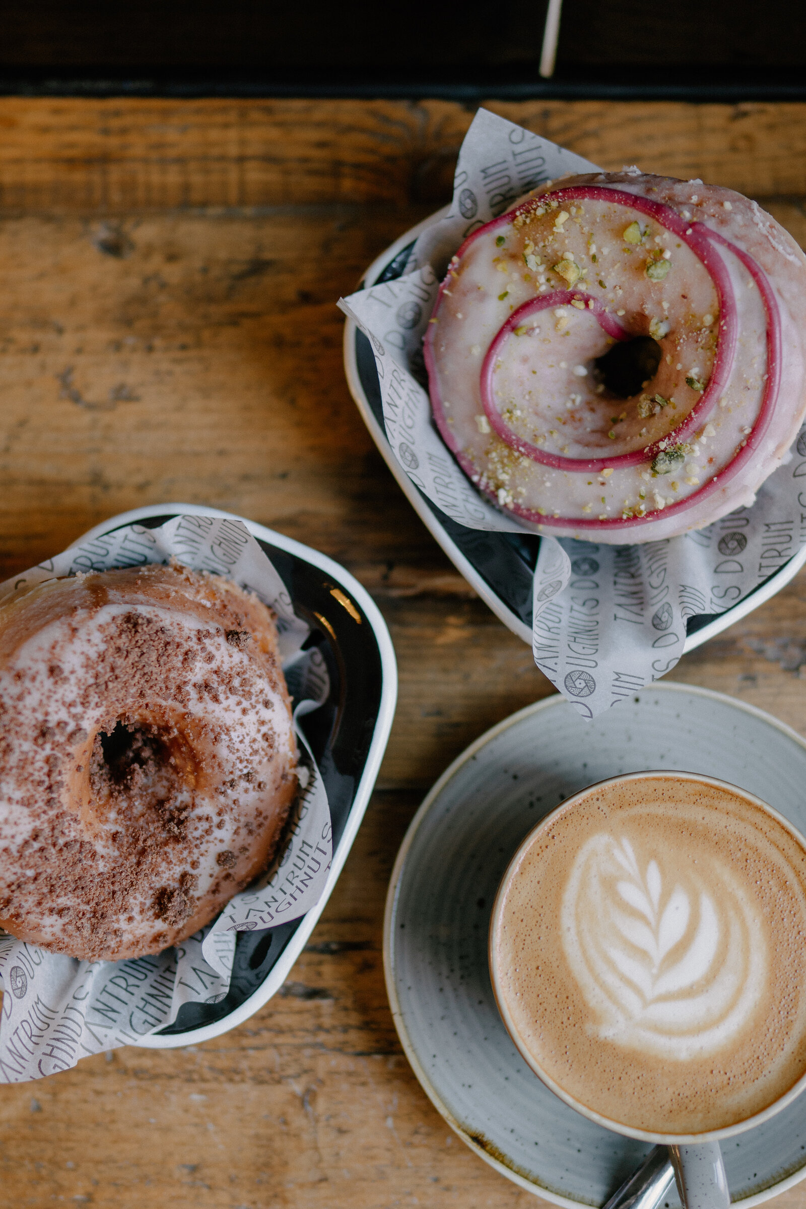 Two glazed doughnuts sit on small plates beside a latte with leaf-pattern foam on a wooden table.