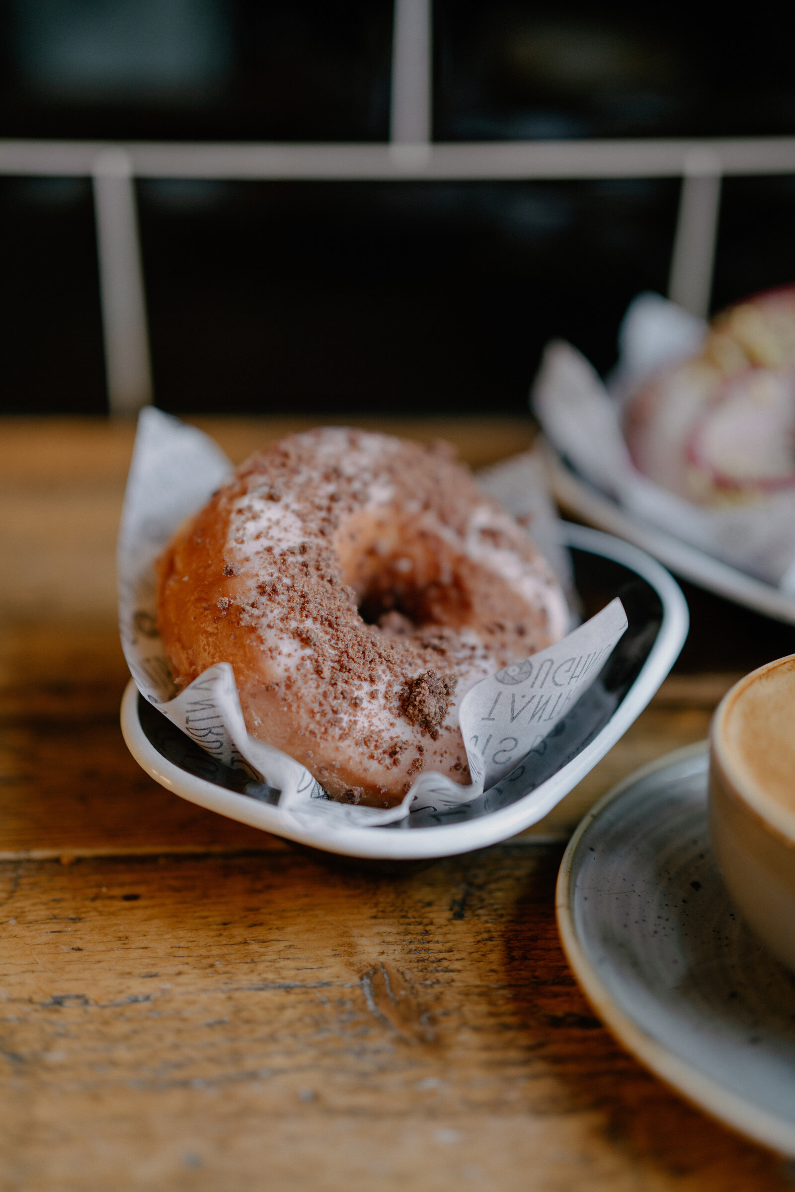 A chocolate-dusted glazed doughnut sits on a plate beside a cup of coffee on a wooden table, another doughnut visible behind.