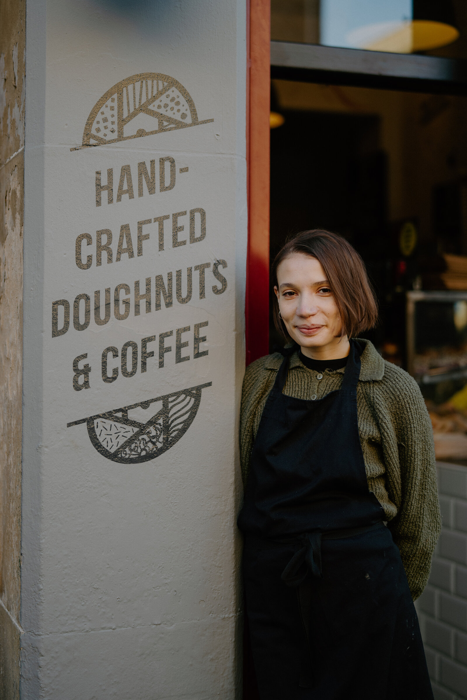 A young woman in an apron stands smiling beside a sign reading 'Hand-Crafted Doughnuts & Coffee' outside a shopfront.