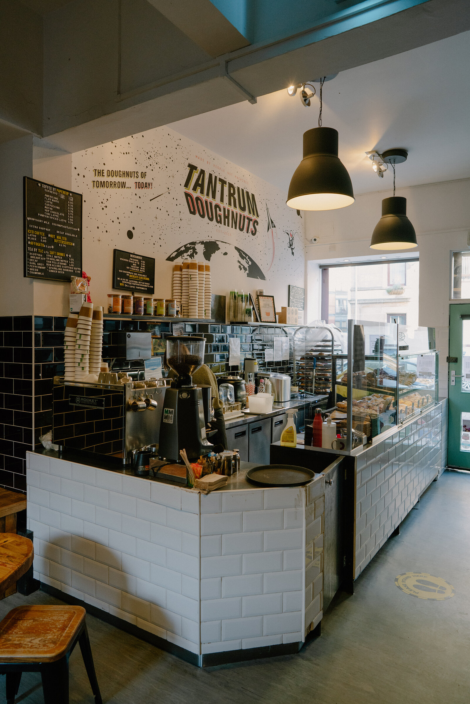 A tiled café counter with a coffee machine and pastry display sits beneath a mural reading 'Tantrum Doughnuts, The Doughnuts of Tomorrow, Today!', hanging pendant lights above.