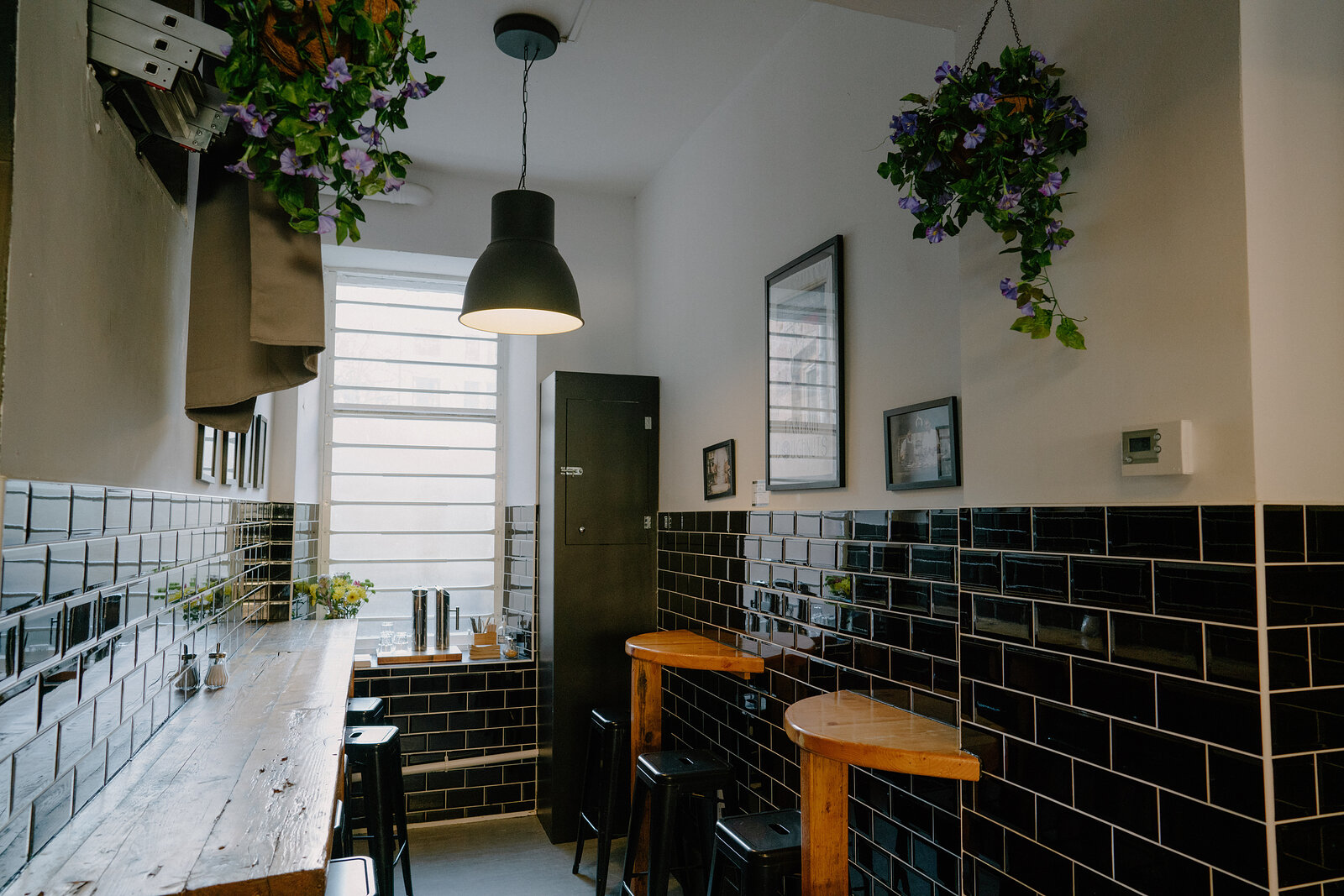 A narrow café corner with black-tiled walls and hanging flower baskets has a wooden bar and stools beneath a pendant light.
