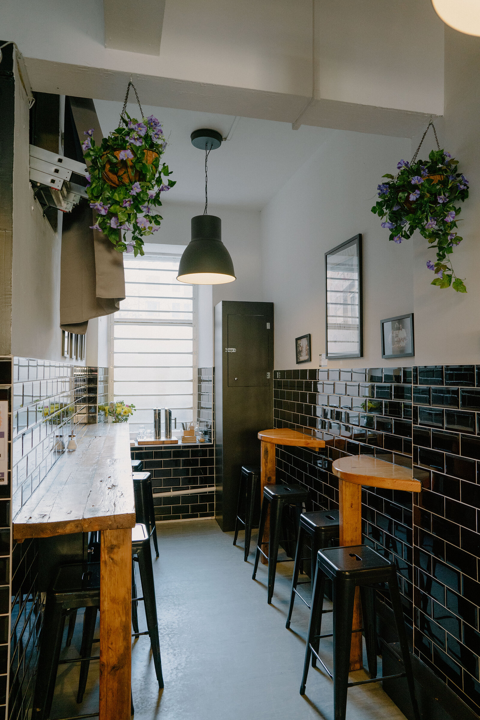 A narrow café corner with black-tiled walls and hanging flower baskets has wooden bar tables and black stools beneath a pendant light, framed pictures on the wall.