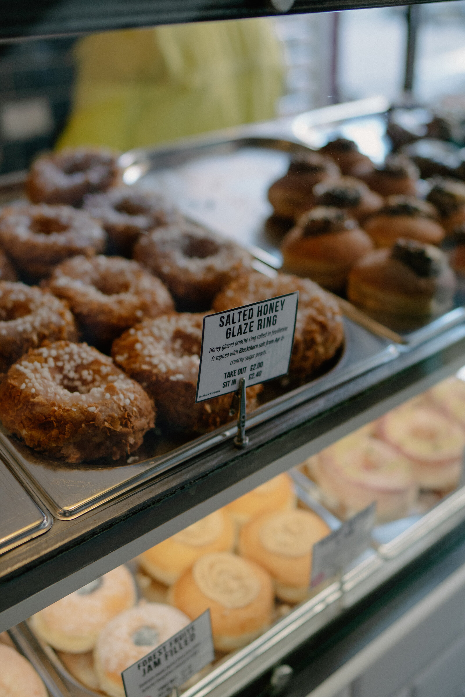 A glass display case shows a tray of salted honey glaze rings labelled with a price card, chocolate-topped pastries and jam-filled doughnuts on the shelves below.