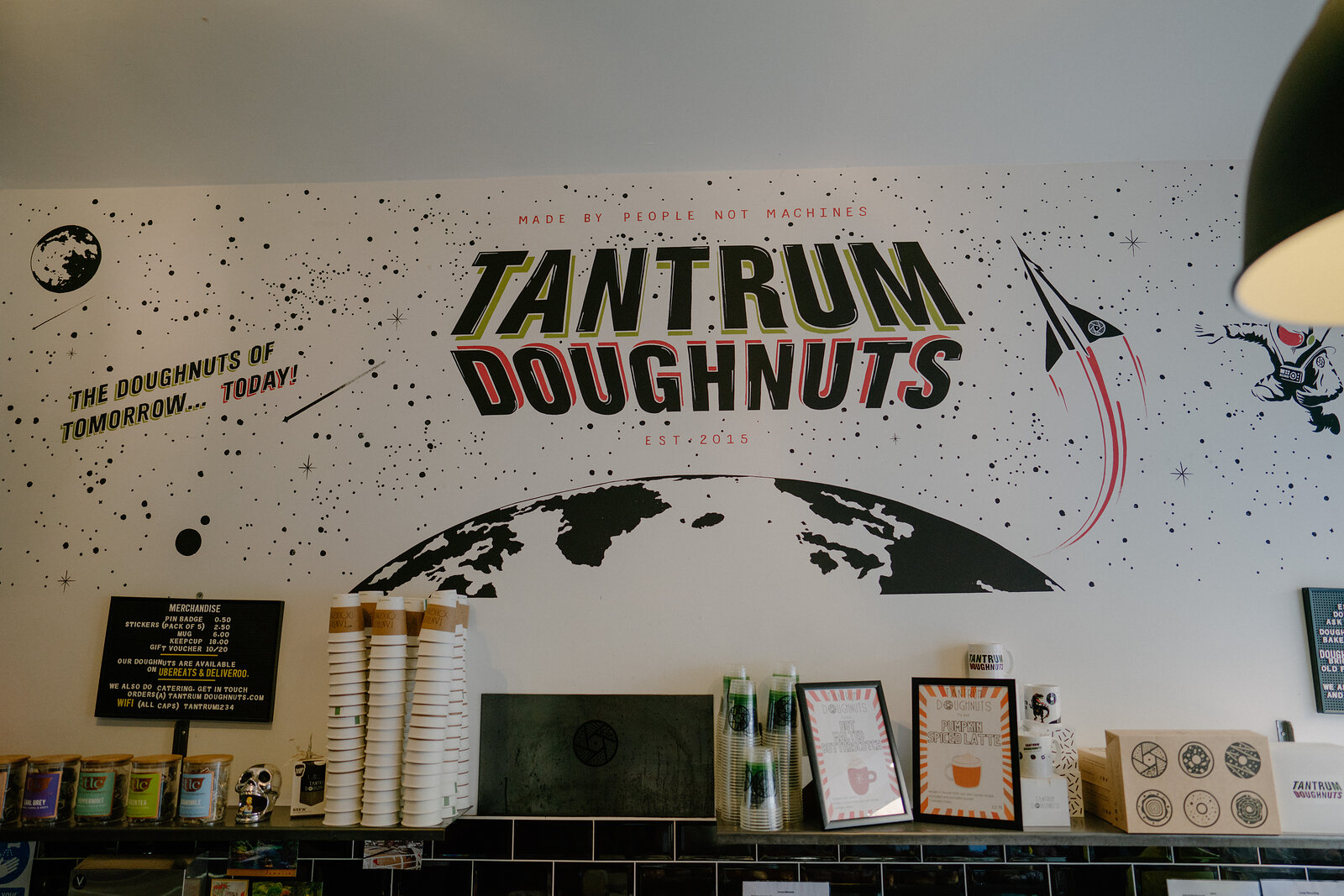 A shop wall painted with a large globe and the words "Tantrum Doughnuts, the doughnuts of tomorrow today, made by people not machines", above a counter with stacked cups and framed artwork.