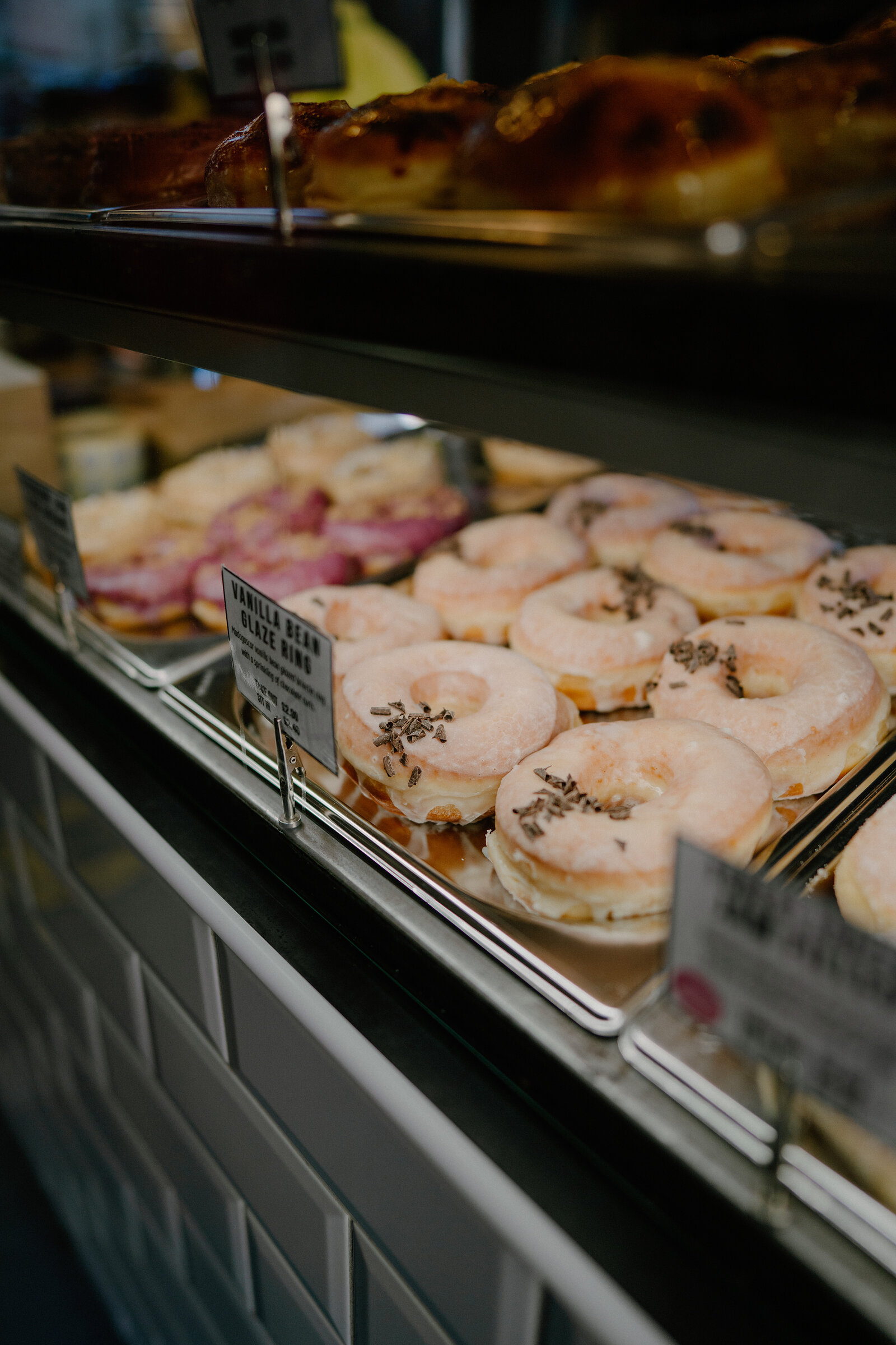 A bakery display case holds trays of iced doughnuts and pastries, with a handwritten label for vanilla bean glaze doughnuts propped in front of a row topped with sprinkles.