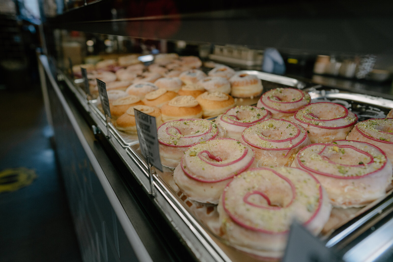 A bakery display case holds trays of glazed rings and open pastries topped with pink onion and herb garnish beside plain iced doughnuts.