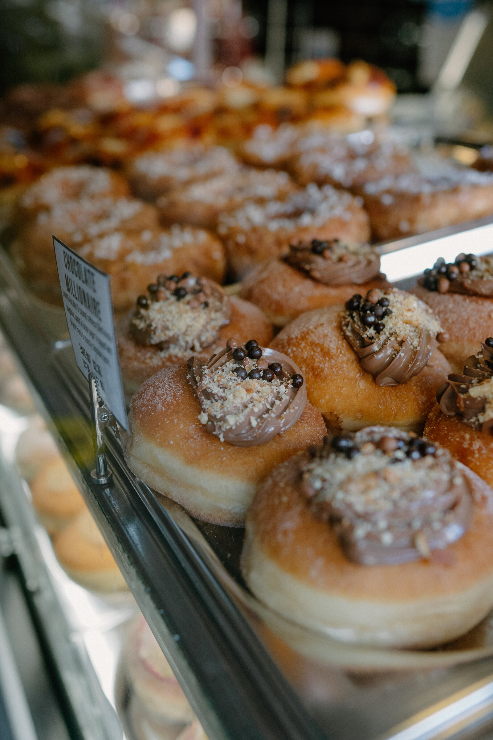 Sugared doughnuts topped with piped chocolate, chopped nuts and gold-flecked sprinkles are arranged in a bakery display case beside a handwritten flavour label.