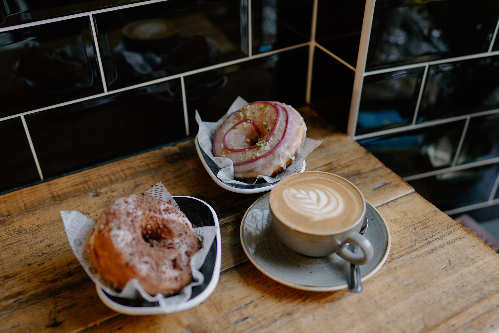 Two glazed doughnuts on paper liners sit beside a flat white with leaf-pattern latte art in a grey mug, on a wooden table against black tiled wall.
