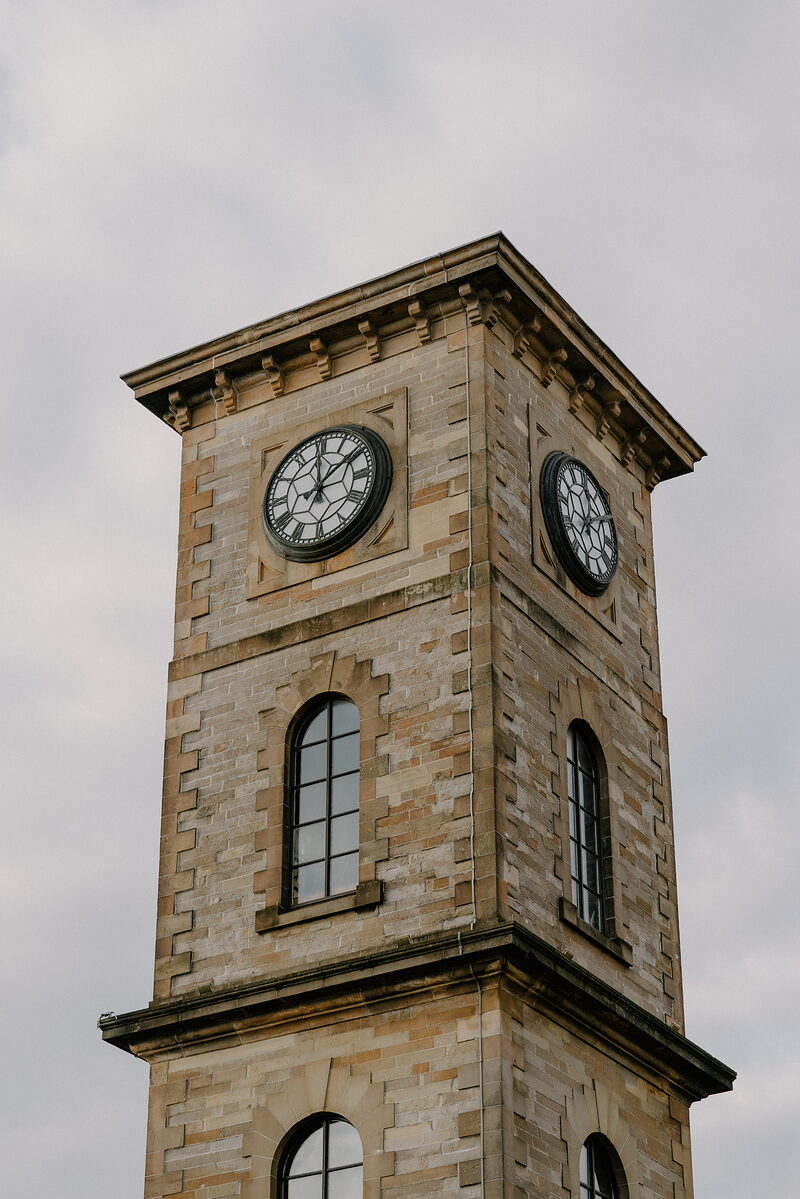 A sandstone clock tower with paired clock faces rises above arched windows on each floor, corbelled stonework running beneath its parapet.