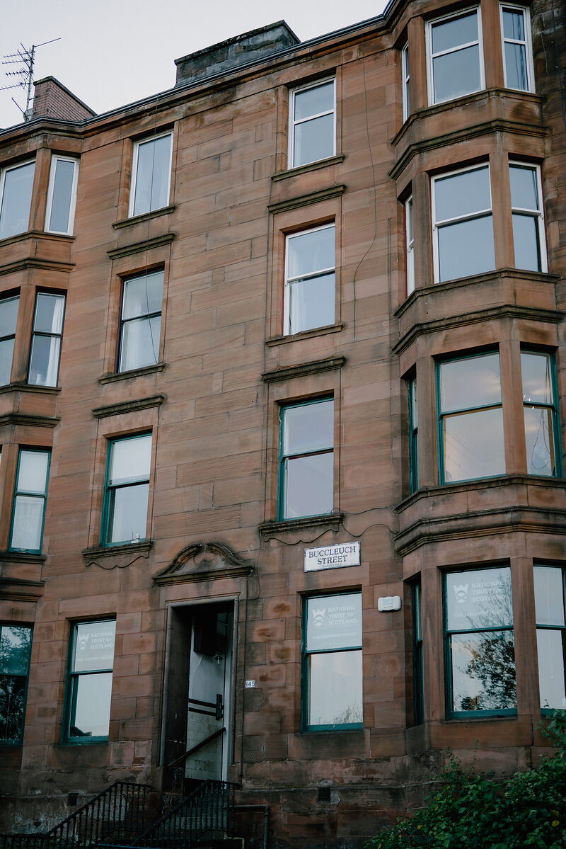 A sandstone tenement building with bay windows and green-framed sash windows rises above a carved doorway lettered "Buccleuch Street".