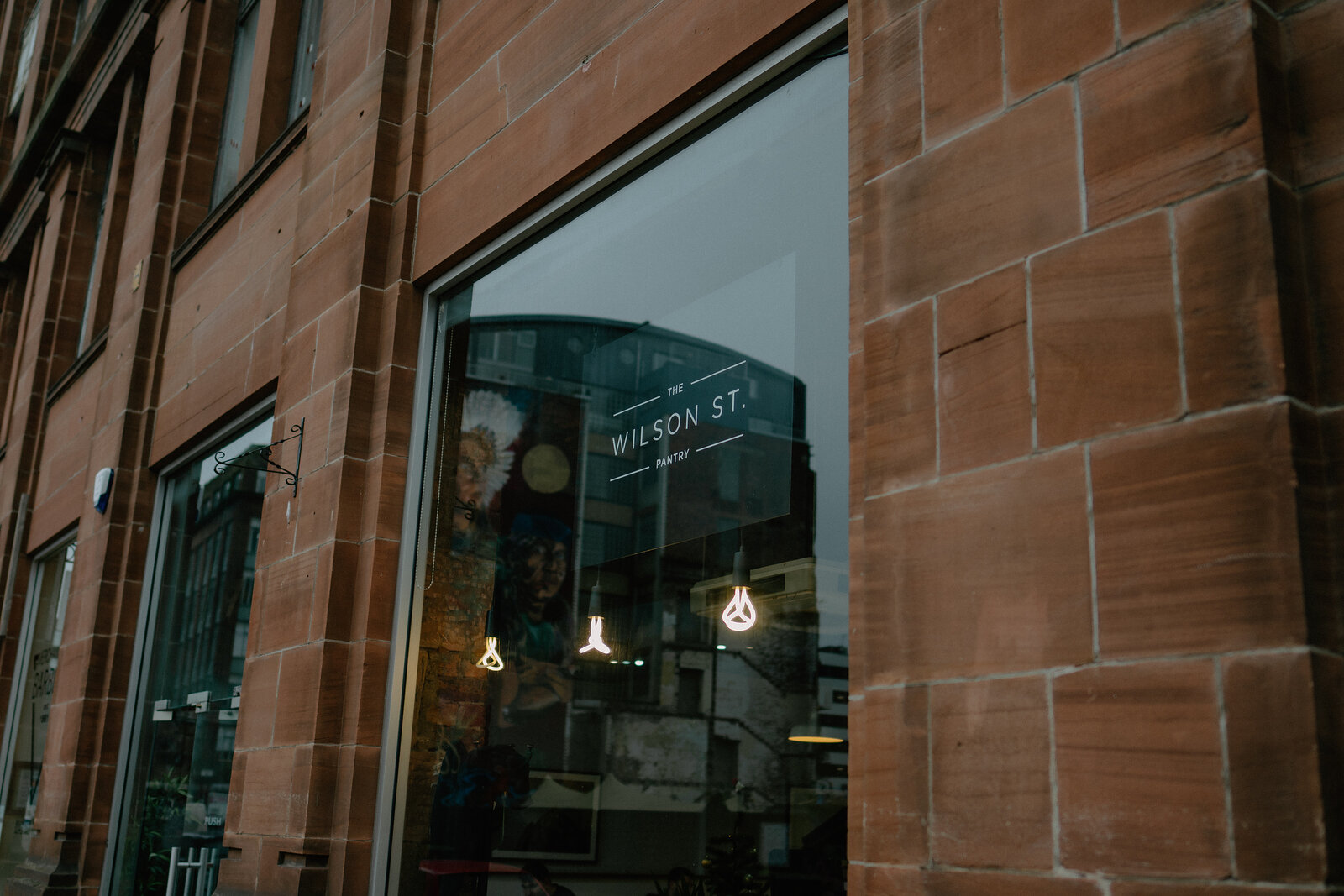A sandstone shopfront window lettered "Wilson St" reflects the street, hanging pendant lights and shelving visible inside.