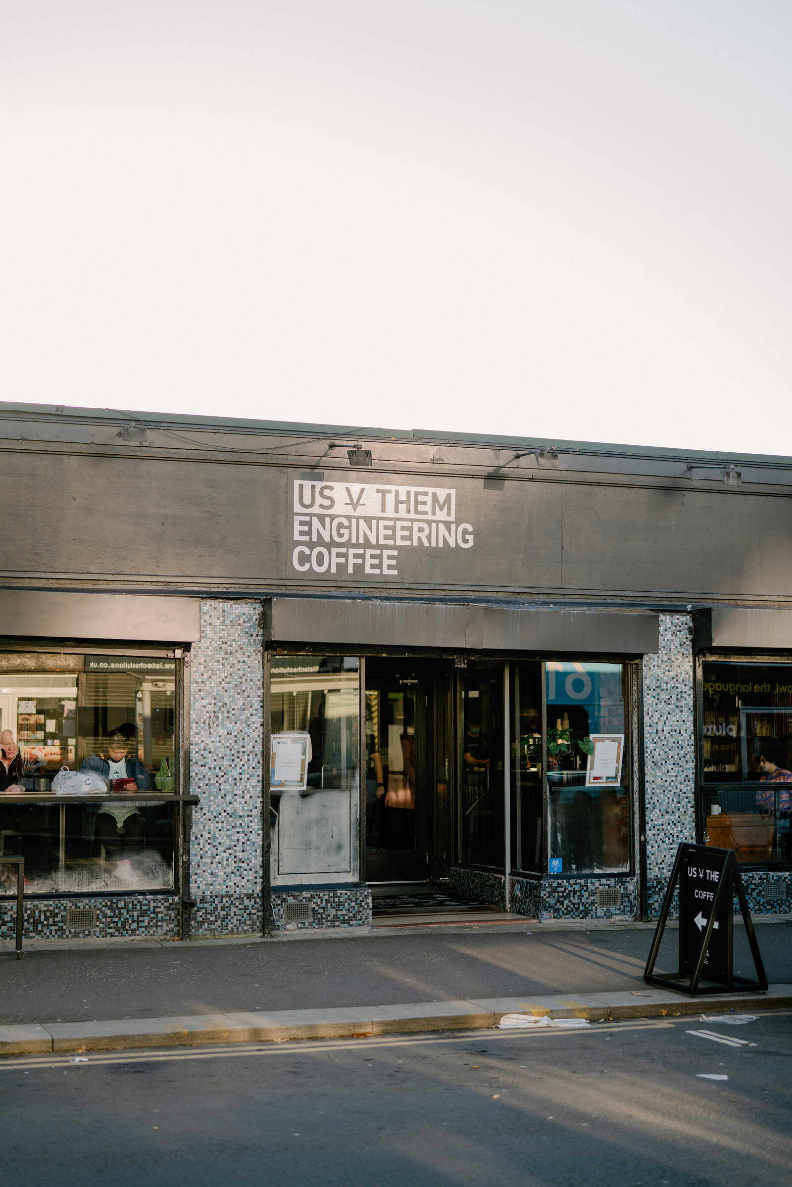 A café shopfront lettered "Us v Them, engineering coffee" has diners seated at a window counter, a sandwich board and outdoor tables on the pavement.