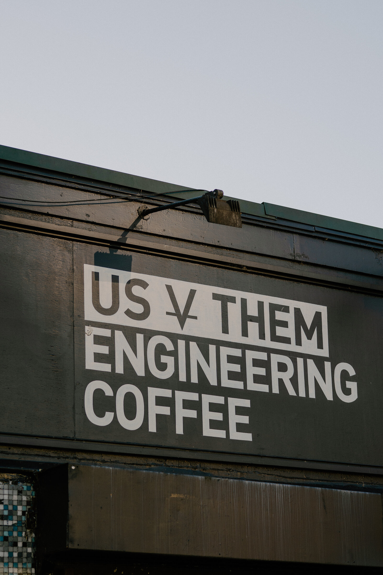 A café building's dark cladding is lettered "Us v Them, engineering coffee" in bold type beneath a wire and light fitting.