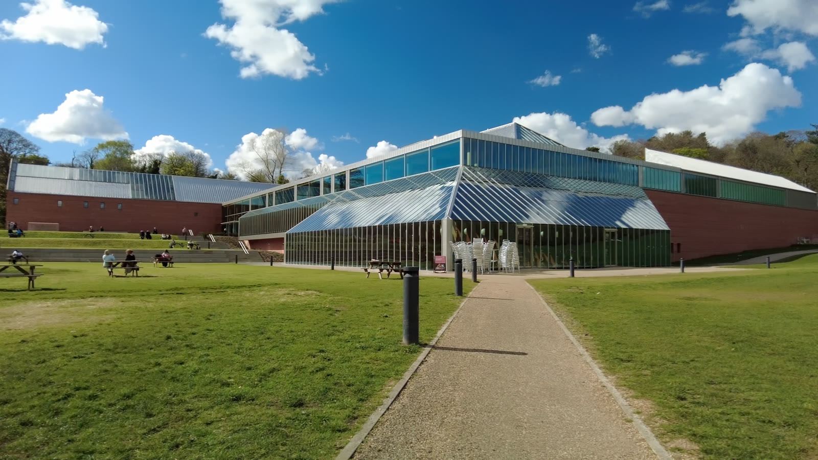 The Burrell Collection across the lawn in Pollok Country Park, its glass-fronted hall between long red sandstone wings, a gravel path leading up to it under a blue sky with white clouds.
