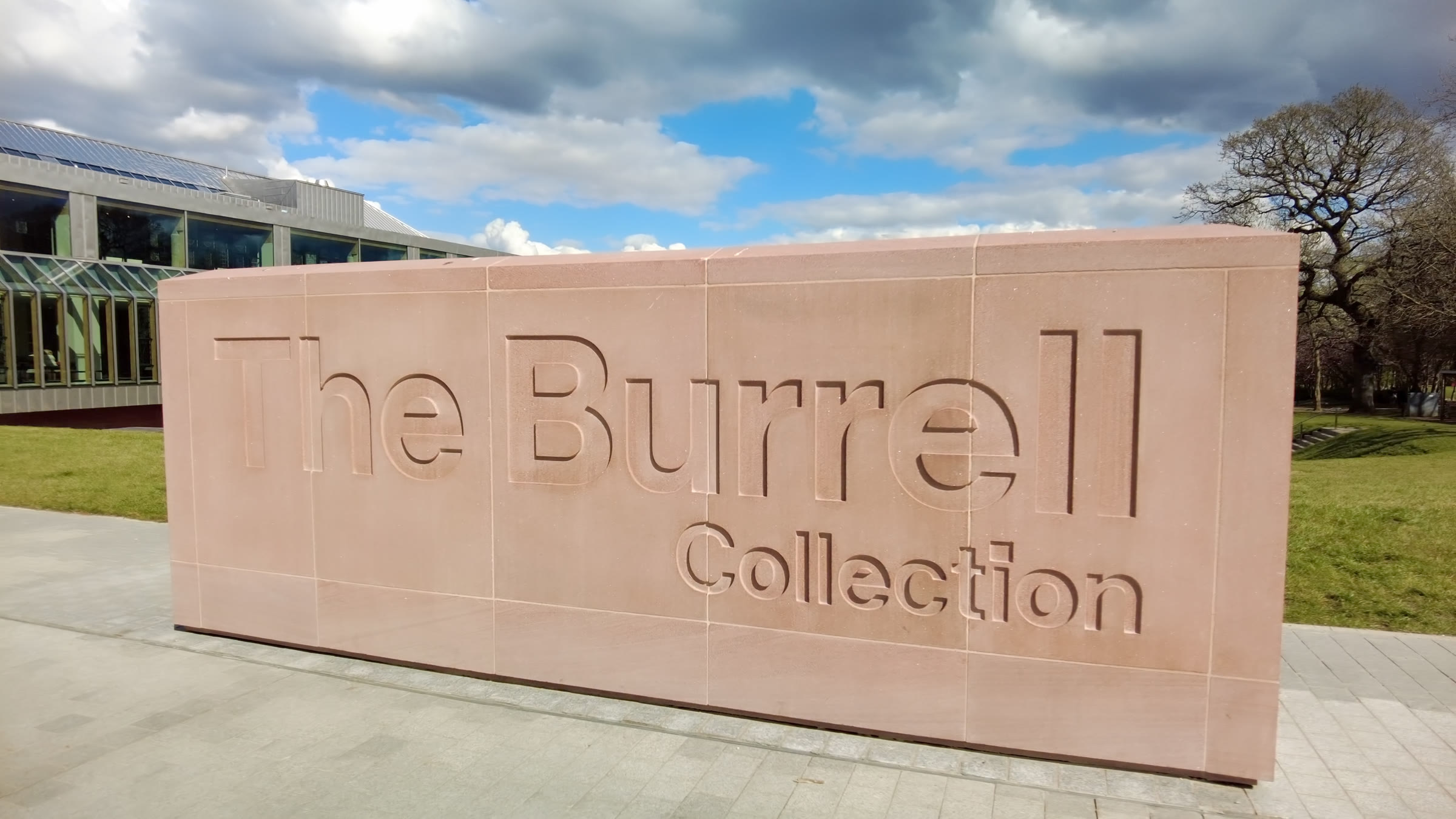 The Burrell Collection's name cut into a long block of pink sandstone at the entrance, the glass and steel front of the building behind it on the left.