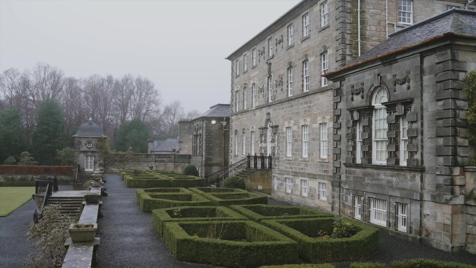 A grand stone mansion's corner tower rises above a box-hedge knot garden, an ornamental summerhouse standing at the end of a gravel terrace.