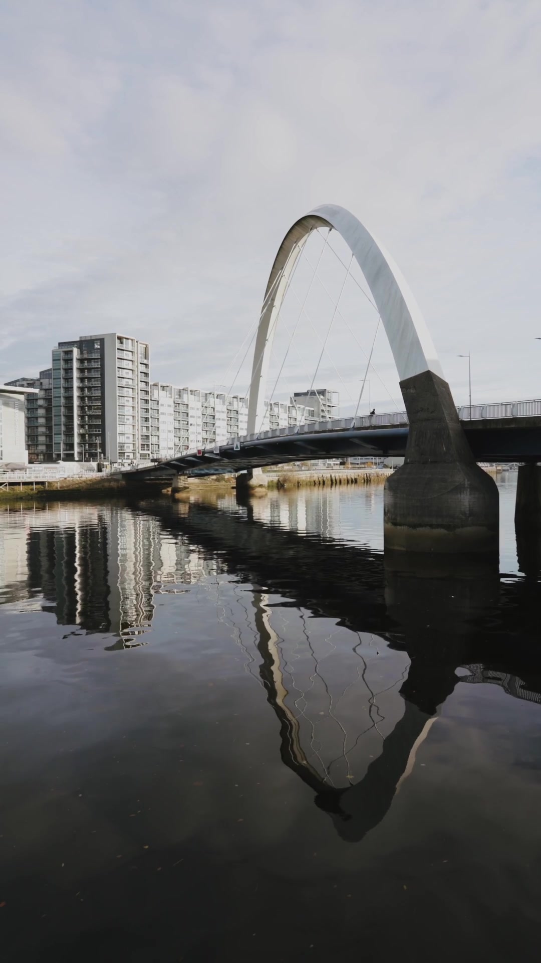 A white arched pedestrian bridge spans a still river between tower blocks, its curve mirrored in the water below.