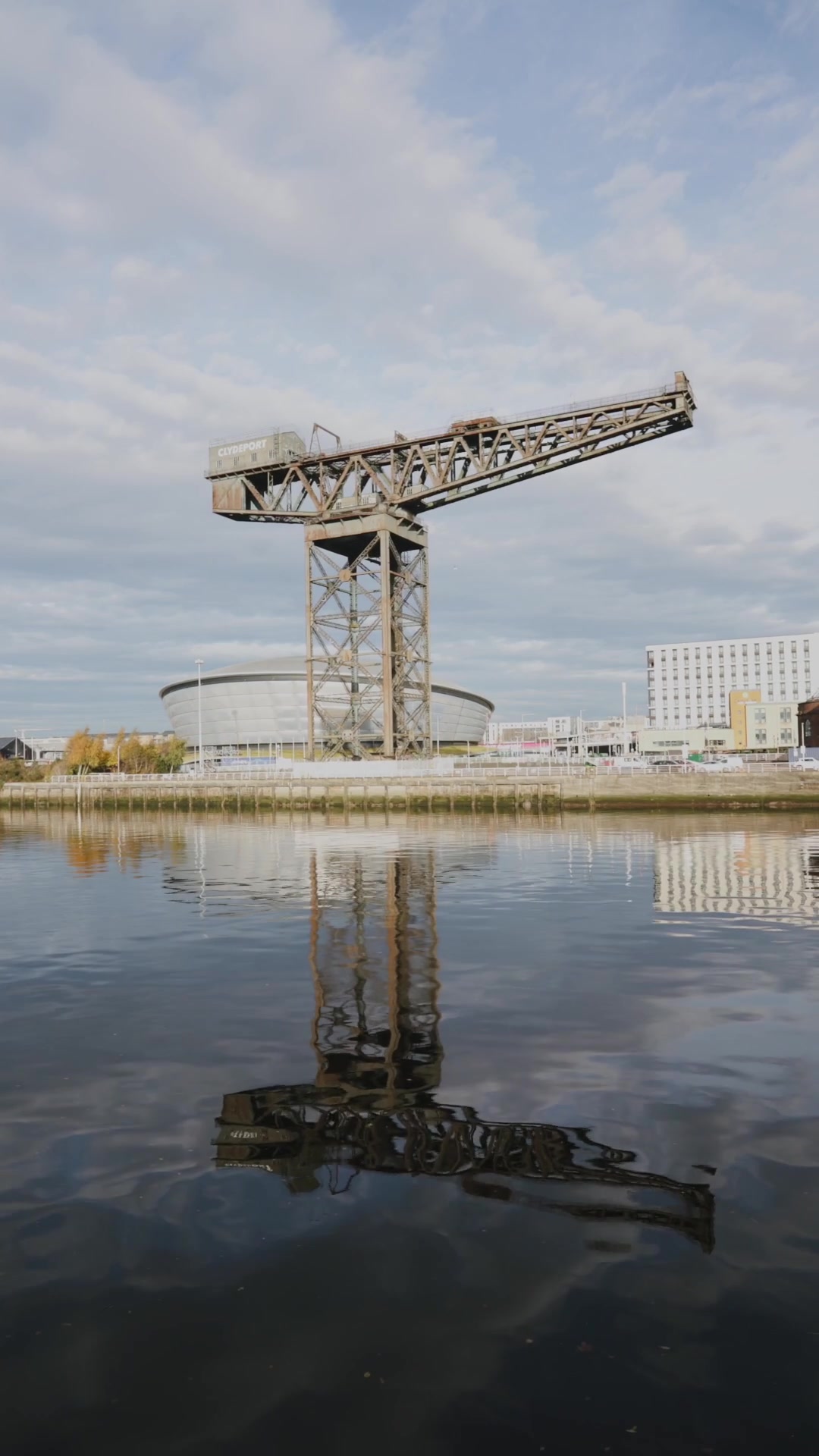 A rusted cantilever crane lettered "Clydeport" stands beside a domed arena, its reflection mirrored in the still river below.