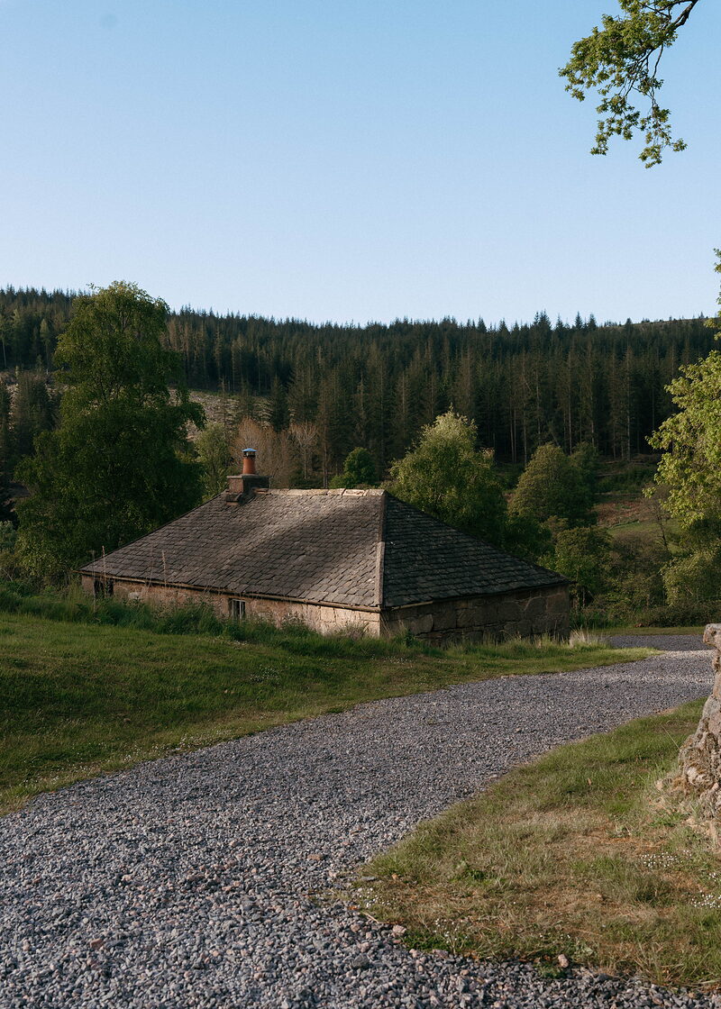 A small stone cottage with a pyramidal slate roof and chimney stands beside a gravel drive, pine forest covering the hillside behind in evening light.