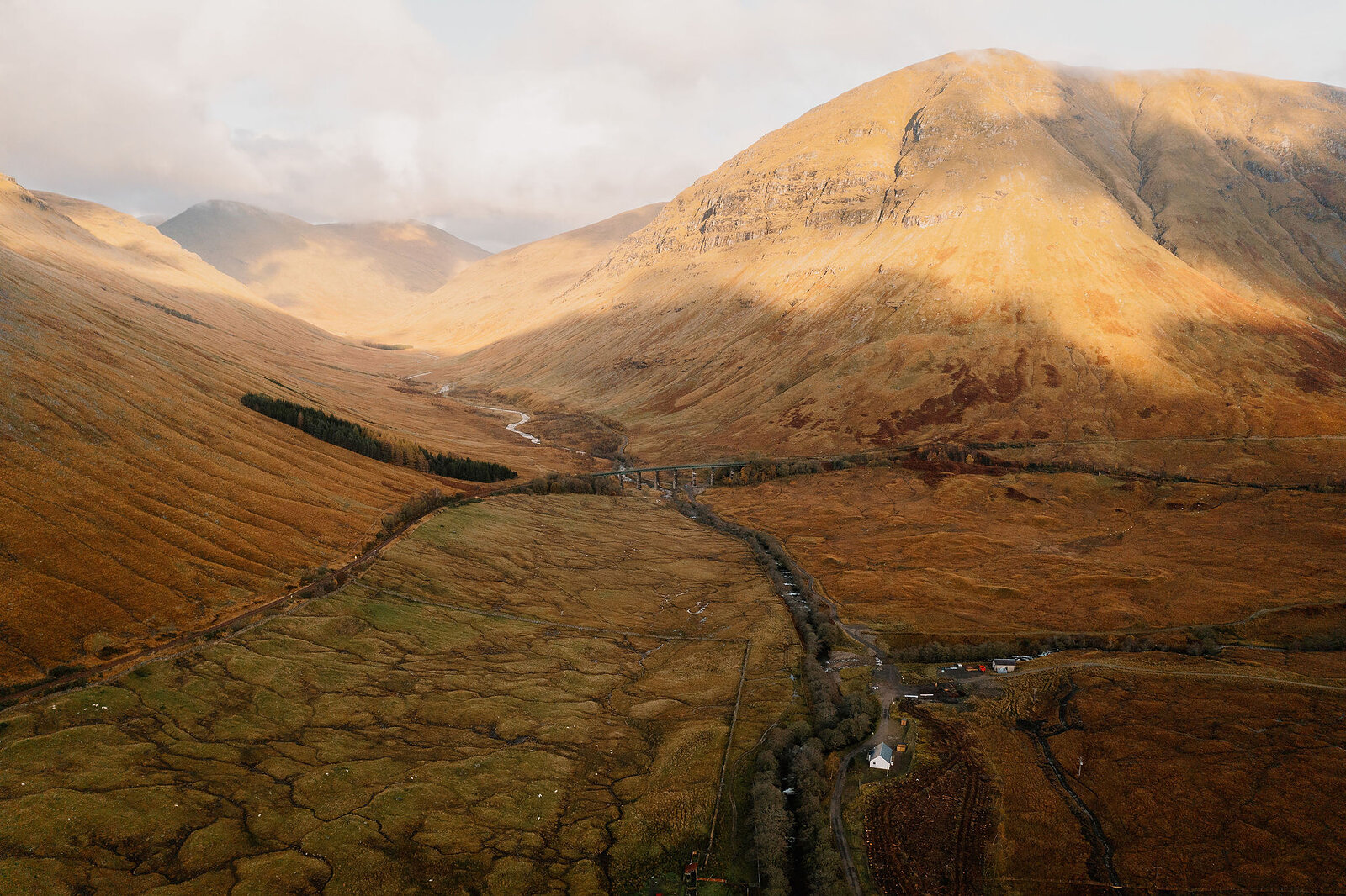 An aerial view of a wide glen shows a road and river running between autumn-coloured hillsides beneath a mountain ridge lit by low sun.