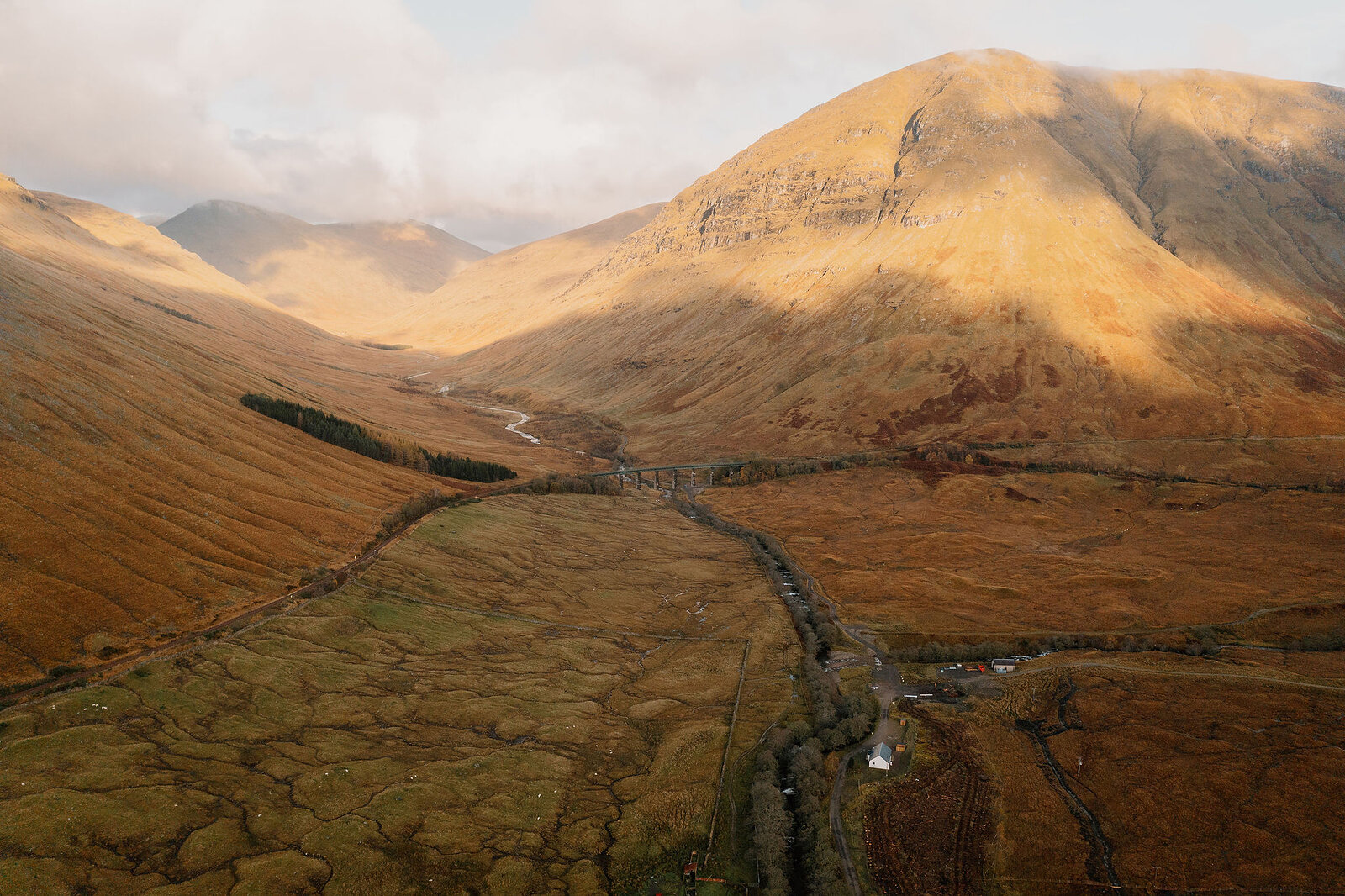 An aerial view of a wide glen shows a road and river running between autumn-coloured hillsides beneath a mountain ridge.