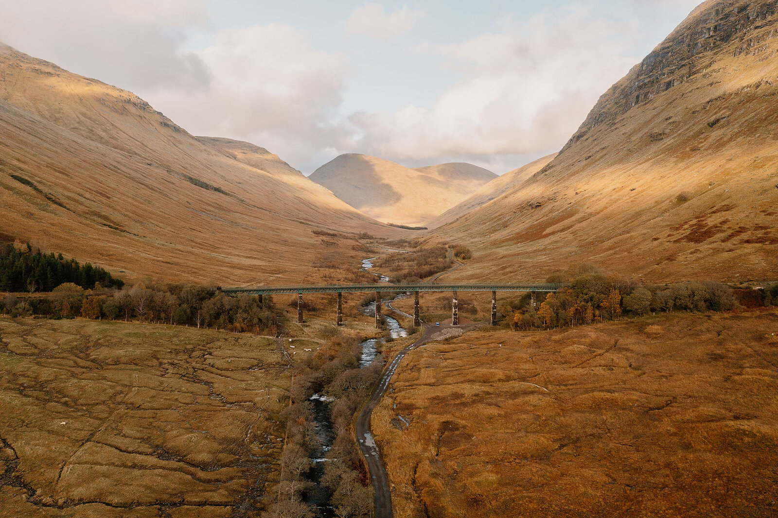 A railway viaduct crosses a river at the foot of a wide glen, autumn-coloured mountains rising steeply on either side.