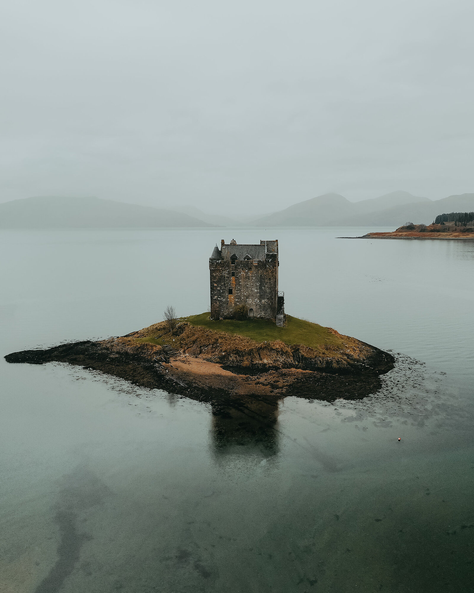A small ruined tower castle stands on a tiny tidal island in a calm loch, mist softening the hills behind and the castle reflected in the water.