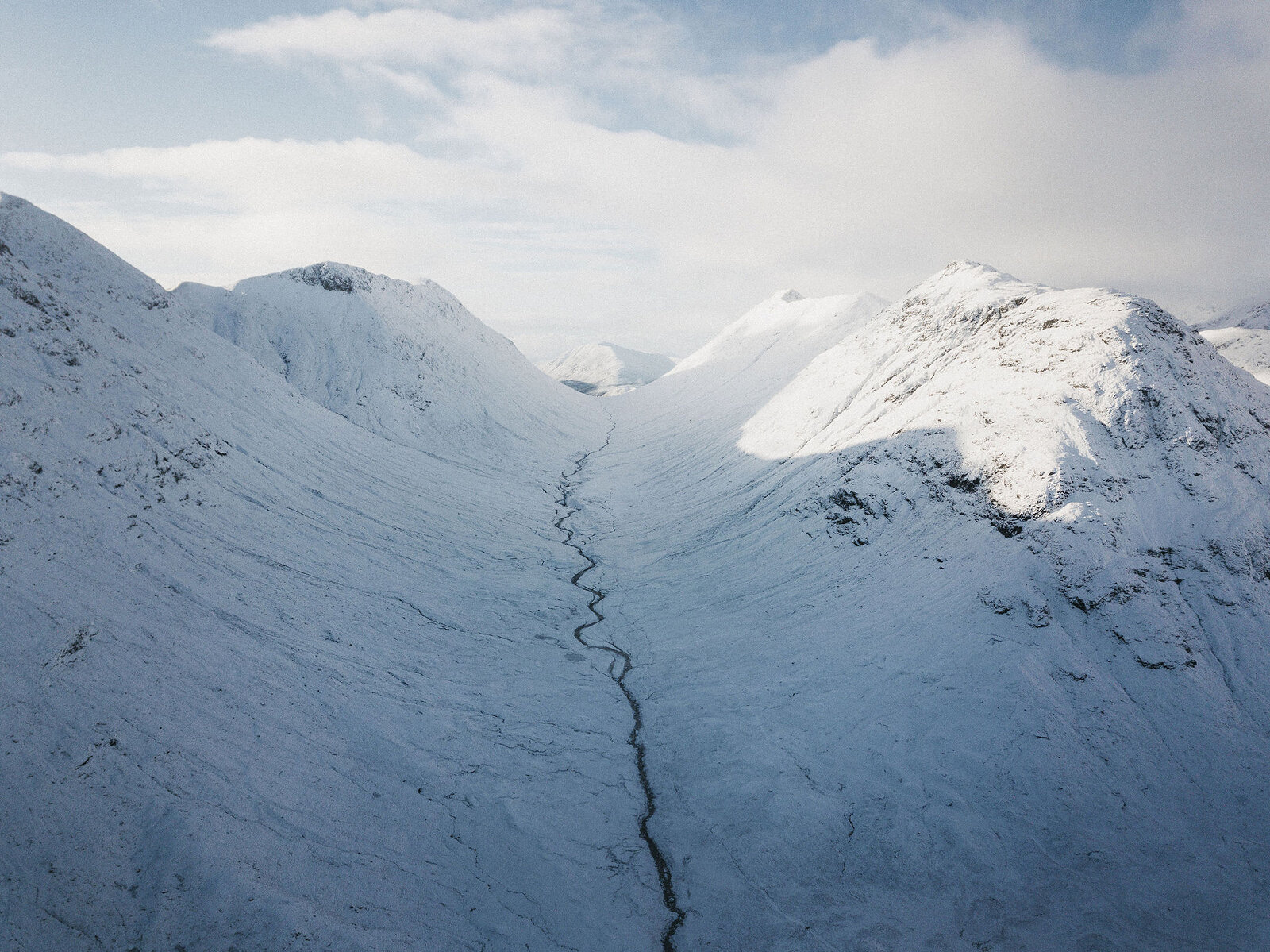 An aerial view shows a snow-covered mountain valley with a stream cutting a dark line through the white slopes.
