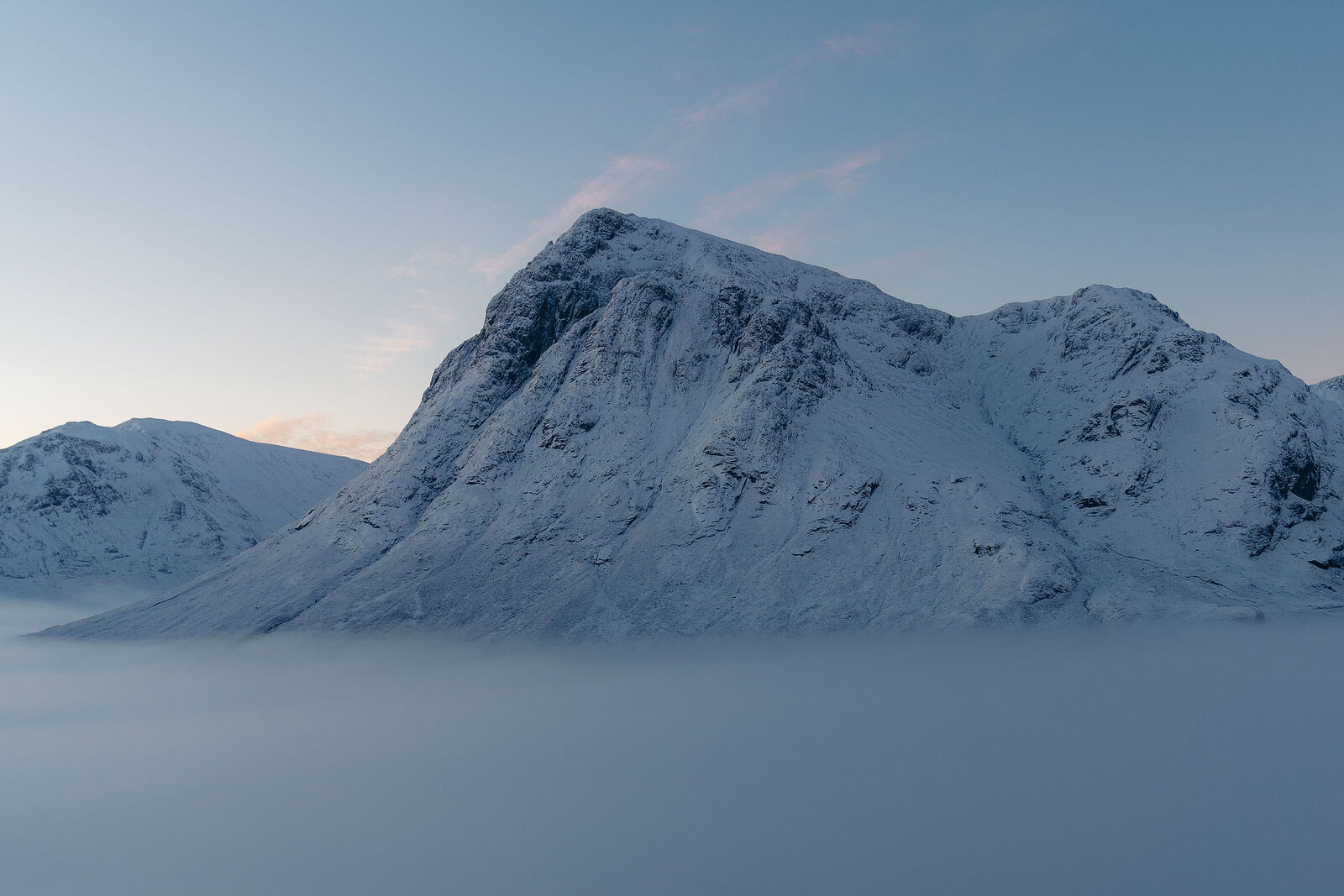 A snow-covered conical mountain peak rises above a layer of mist, pale sky glowing behind it.