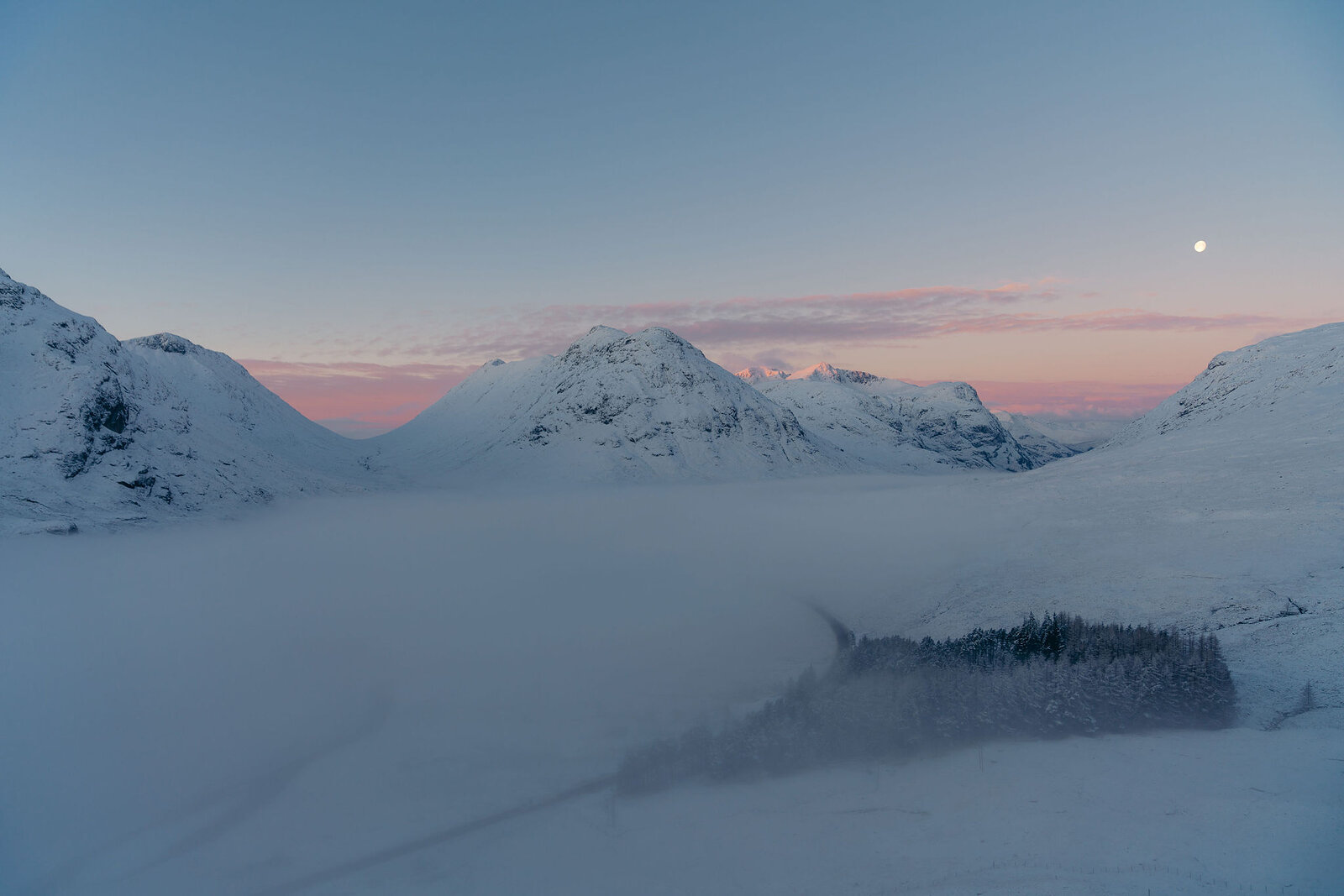 Snow-covered mountains rise above a mist-filled glen at dusk, a pale moon visible in a sky streaked pink and blue.