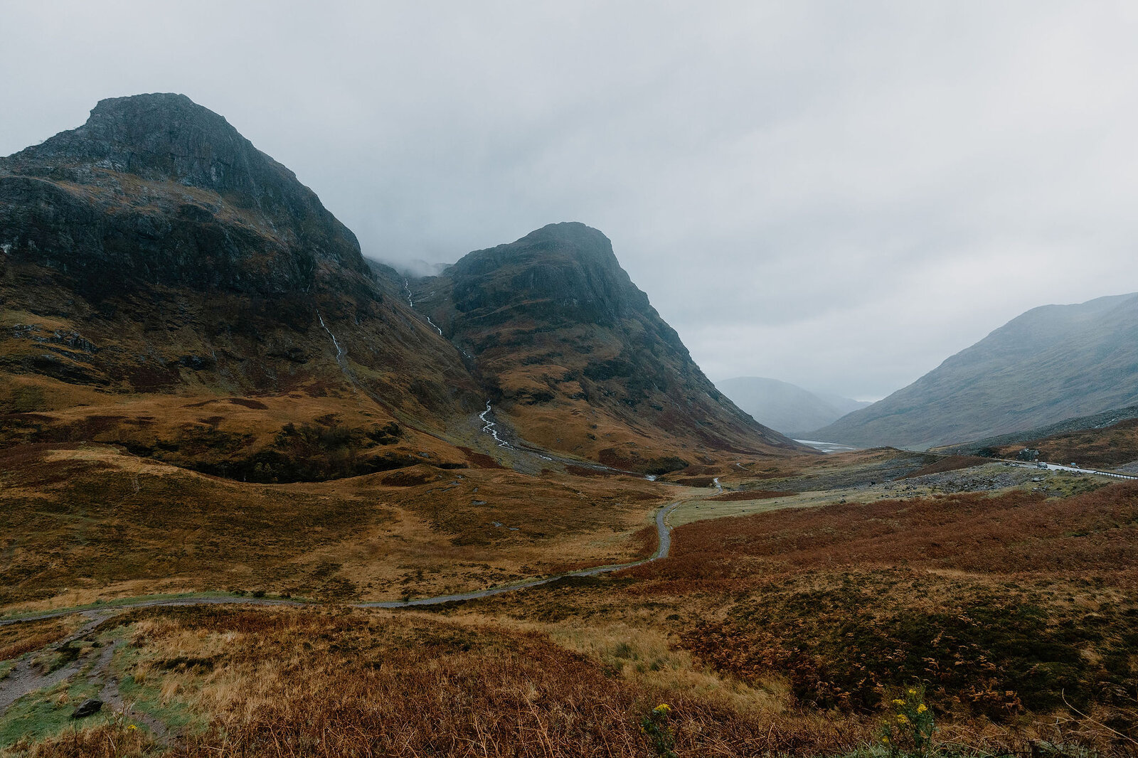 Two rugged mountain peaks rise above a moorland glen, a road and river winding through heather and bracken below.