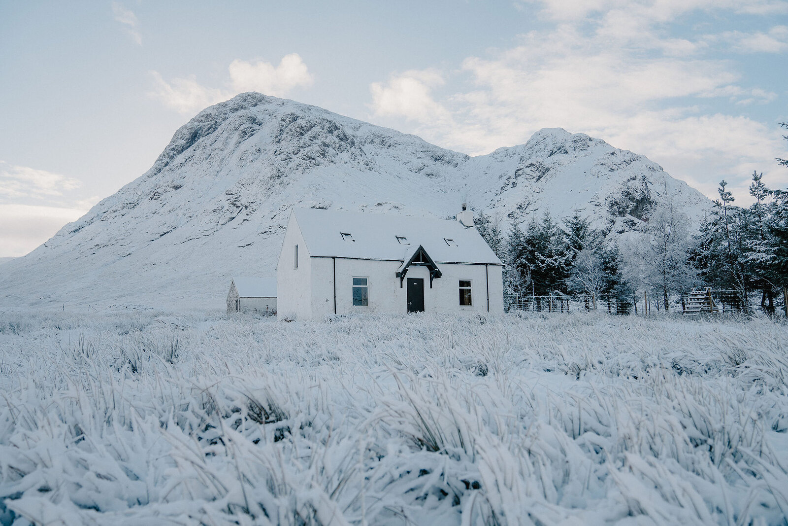 A white cottage stands beneath a snow-covered pyramidal mountain, frosted grass filling the foreground.