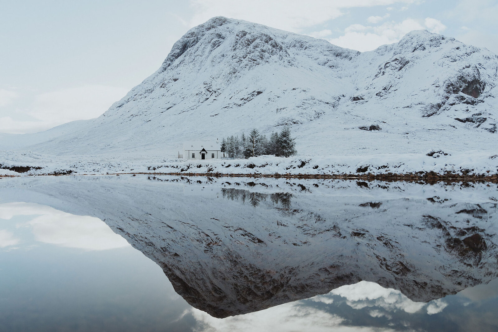 A snow-covered mountain reflects in the still water of a loch, a white cottage standing at the base.
