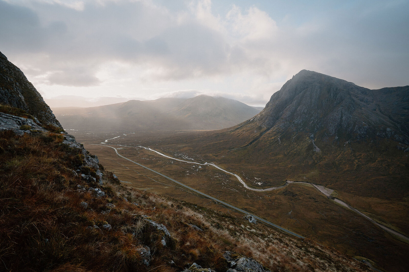 A winding road crosses a misty glen between mountains, low cloud drifting over the peaks.