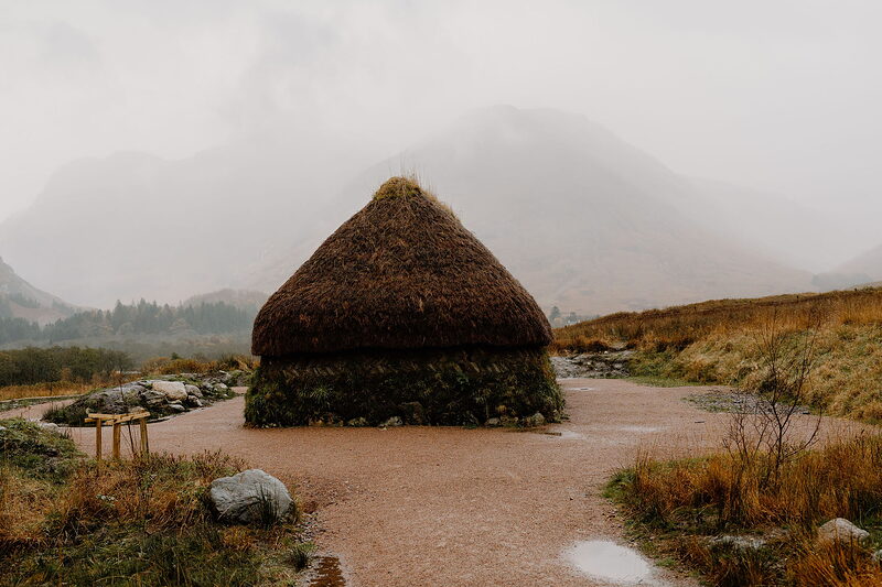 A conical thatched roundhouse stands on a gravel path, mist obscuring the mountains behind.