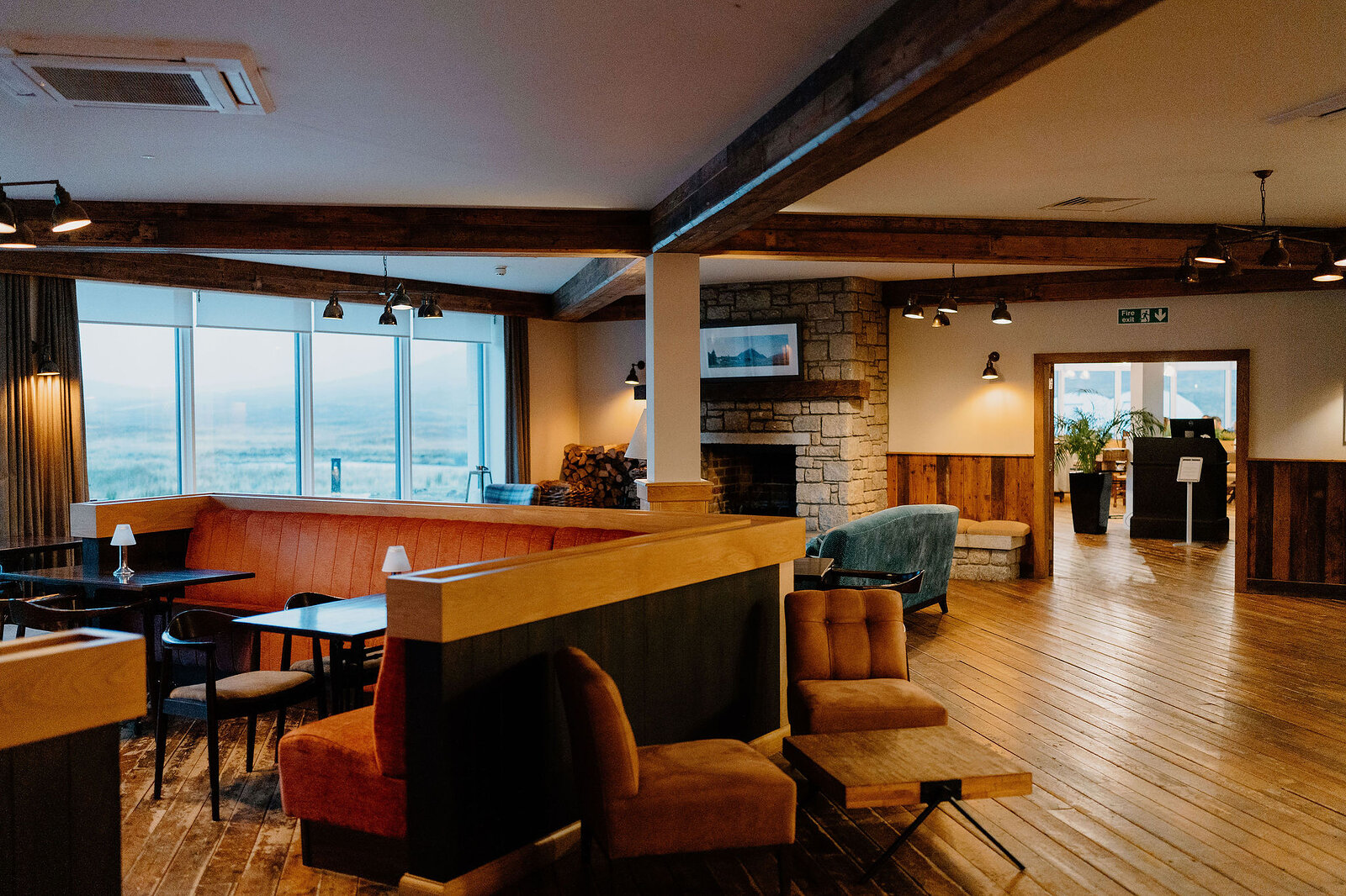Orange armchairs and a sofa surround a stone fireplace in a lounge with a wall of windows overlooking the hills, exposed beams crossing the ceiling.