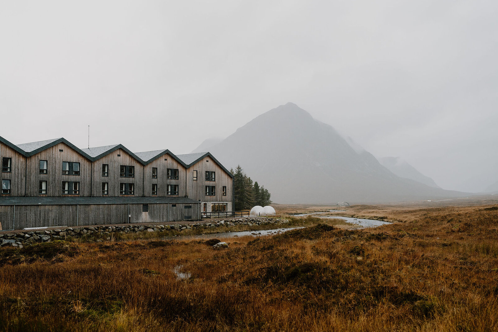 A timber-clad hotel building stands beside a stream and moorland, mist obscuring a mountain behind.