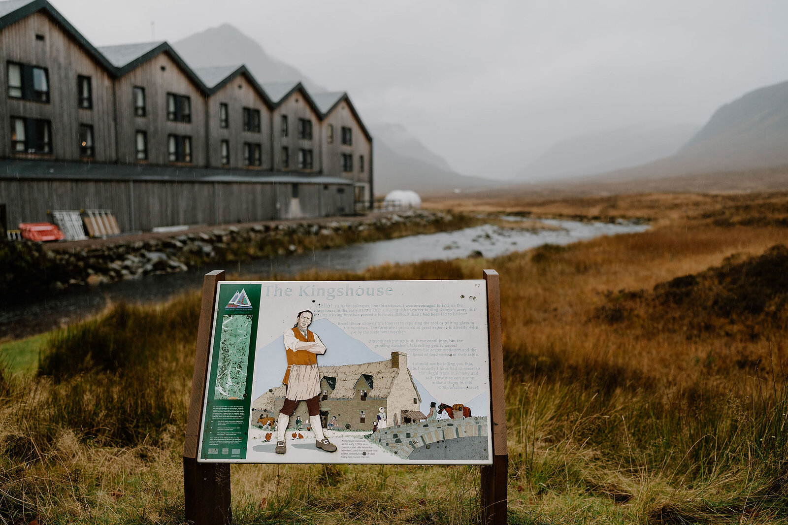 An illustrated interpretive sign about "The Kingshouse" stands before a timber-clad hotel building, a river and misty moorland behind.
