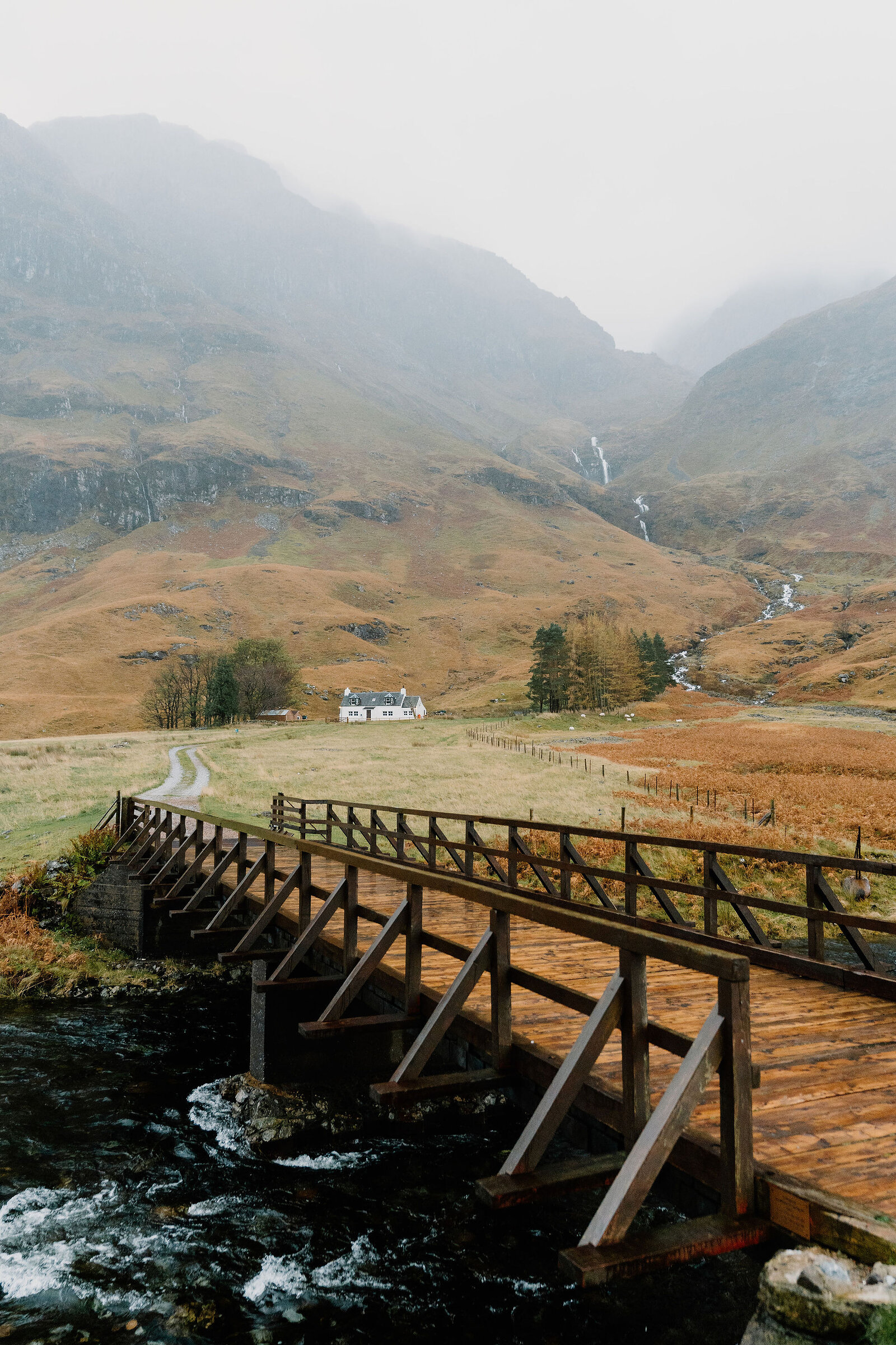 A wooden footbridge crosses a stream leading to a white cottage in a misty glen, waterfalls streaking the mountainside behind.