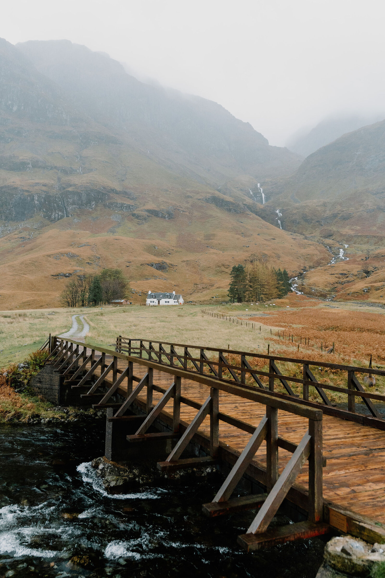 A wooden footbridge crosses a stream leading to a white cottage in a misty glen, waterfalls streaking the mountainside behind.