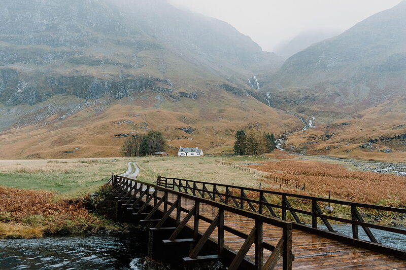 A footbridge over the river to the white cottage of Achnambeithach, the glen rising behind.