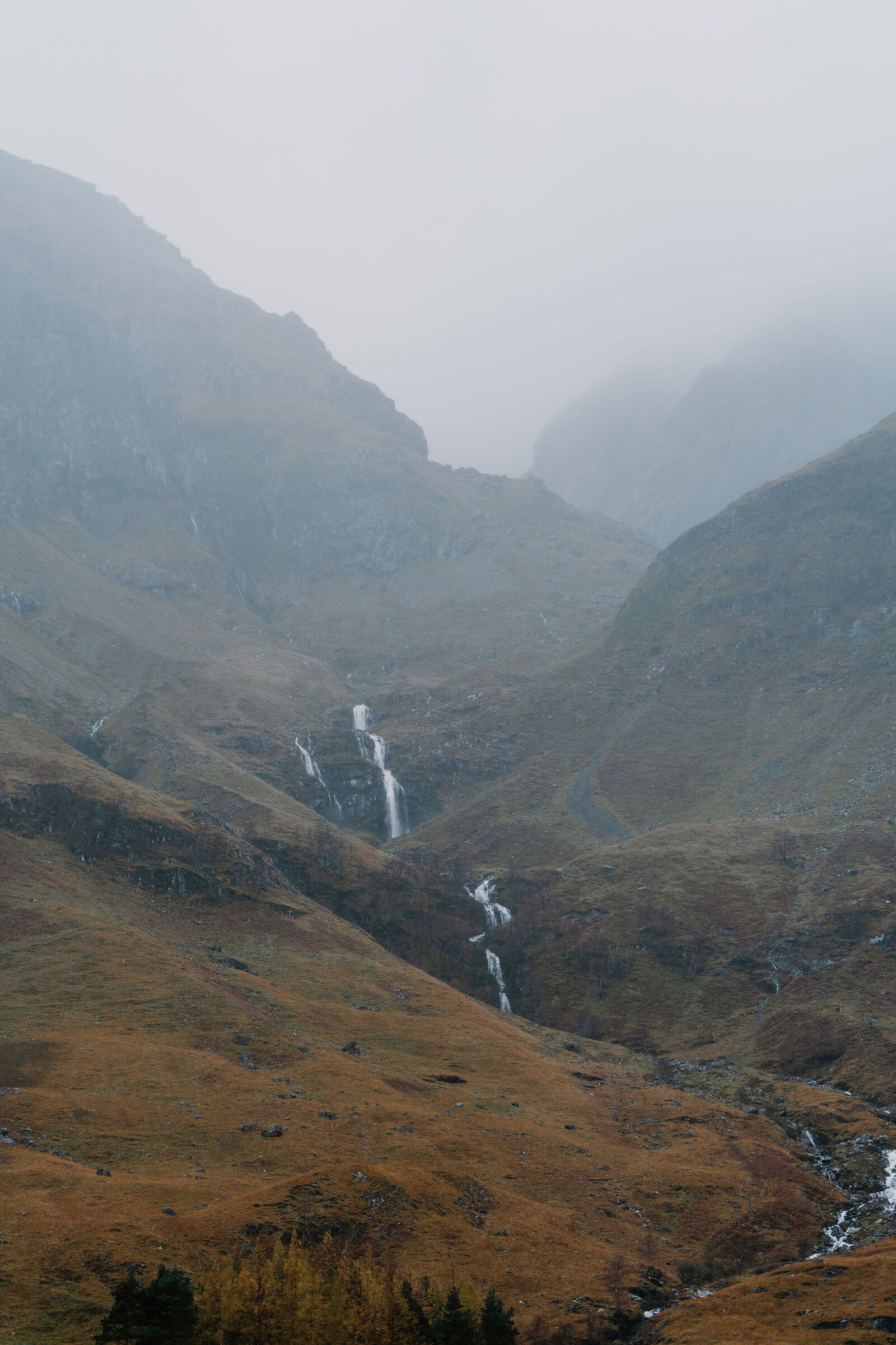 Waterfalls streak down the steep, misty walls of a mountain corrie above autumn-coloured moorland.