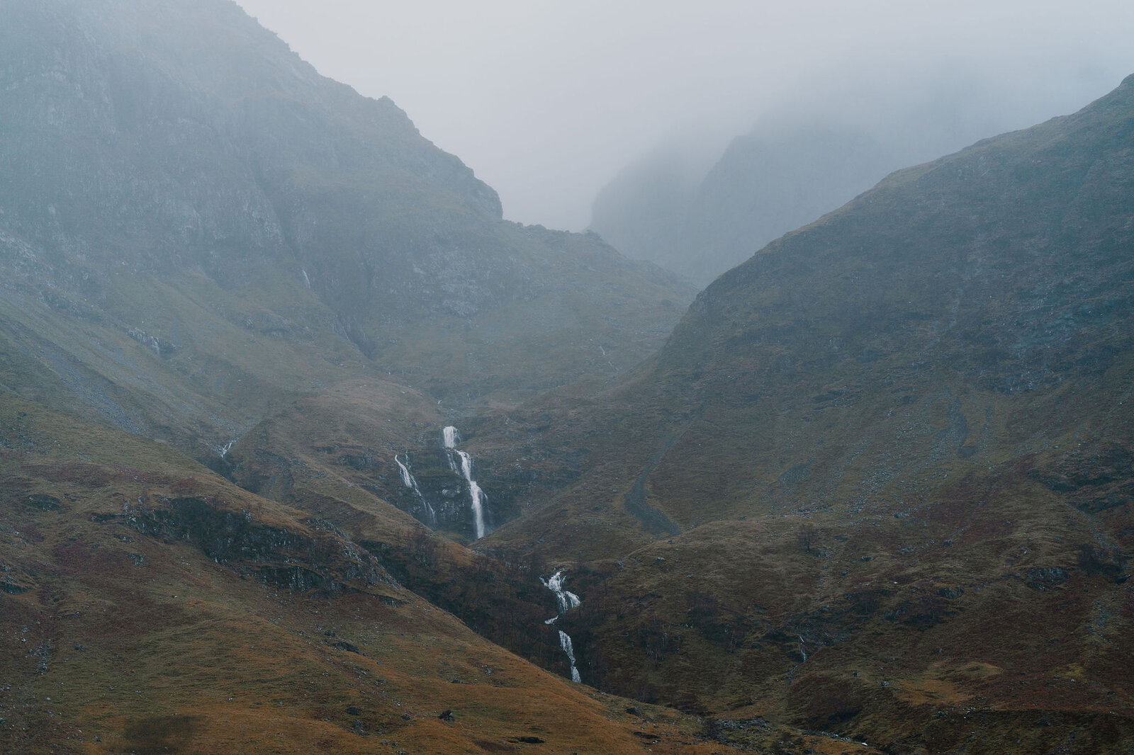 Waterfalls streak down the steep, misty walls of a mountain corrie above autumn-coloured moorland.