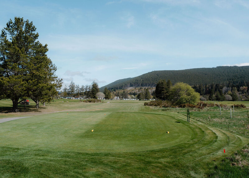 A tee with yellow markers looking down a fairway past pines to white caravans and a forested hill.