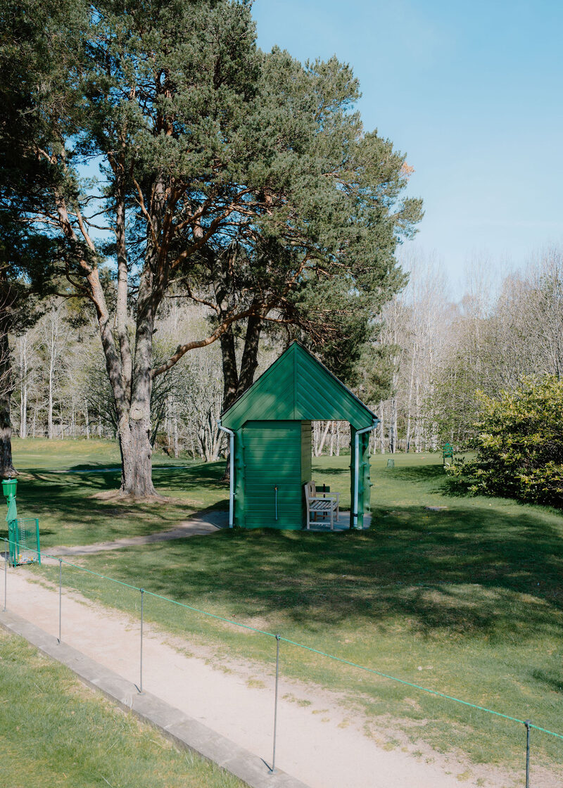 A small green wooden shelter with a bench under Scots pines on the course.